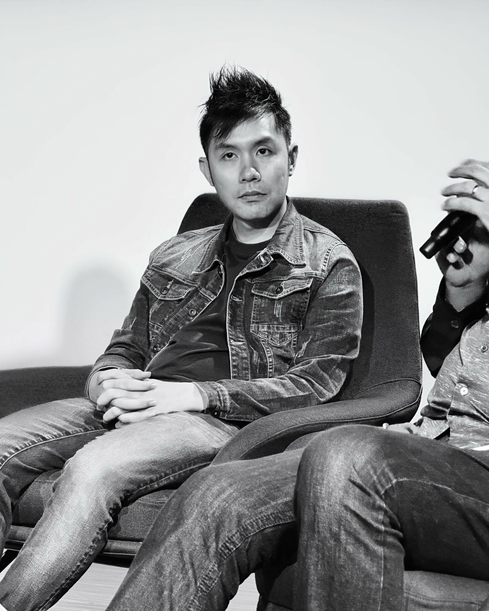 Man sitting on a chair with hands clasped, wearing a denim jacket and jeans, looking at the camera.