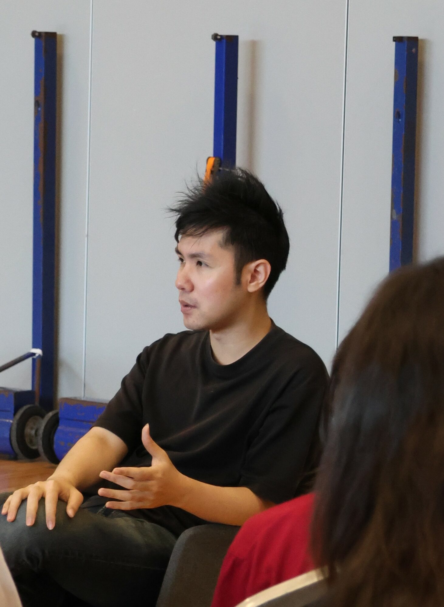 Young man with black hair wearing a black shirt, sitting and gesturing with his hand, in a room with blue and white walls.