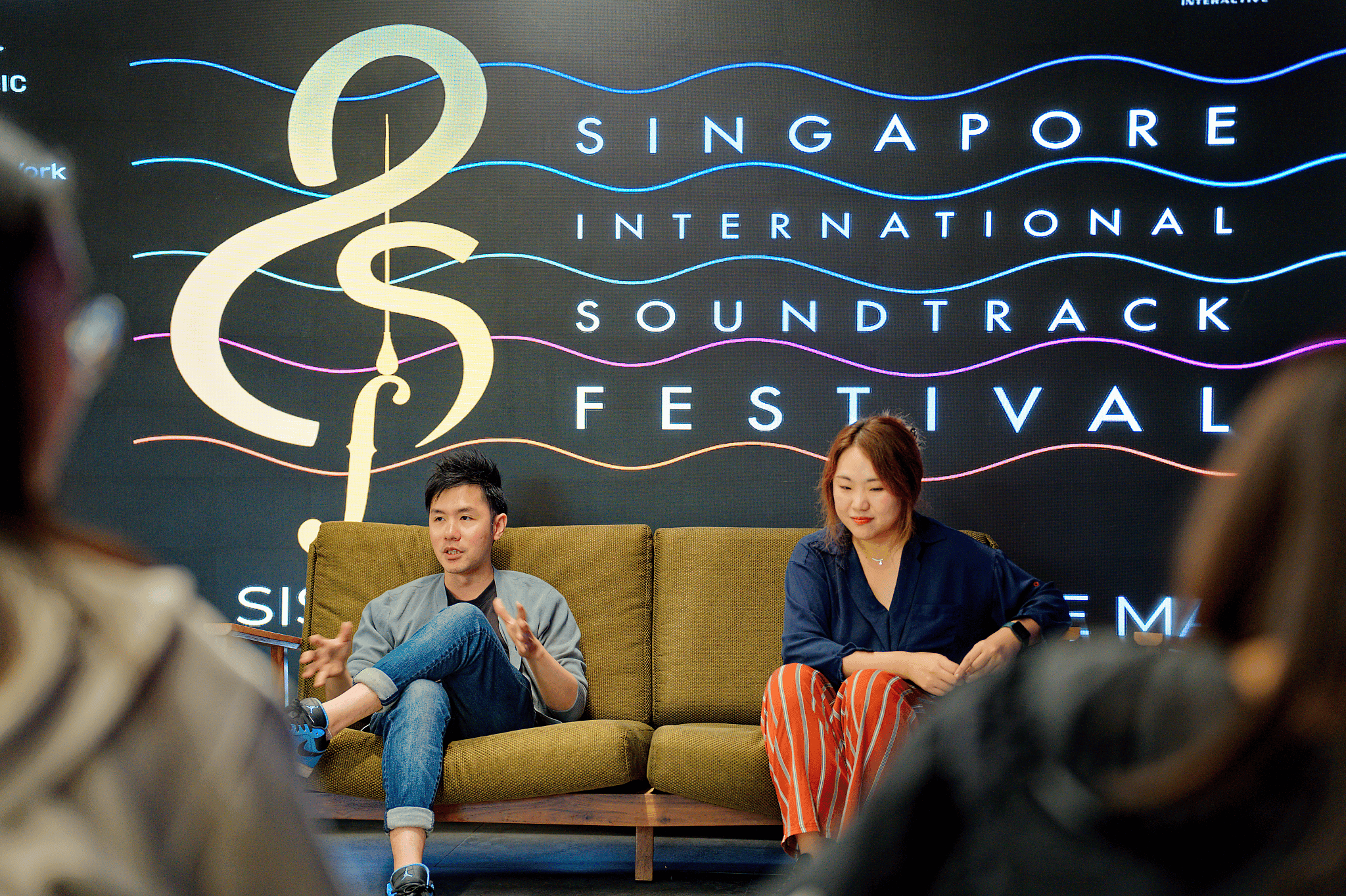 Two people sit on a green sofa in front of a black backdrop with colorful text for the Singapore International Soundtrack Festival, with blurred audience members in foreground.