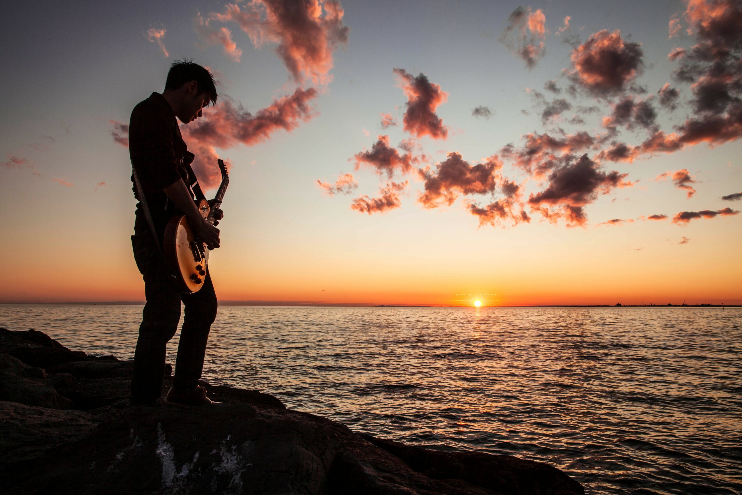 Silhouette of a person playing guitar on rocks by the sea during sunset with clouds in the sky.