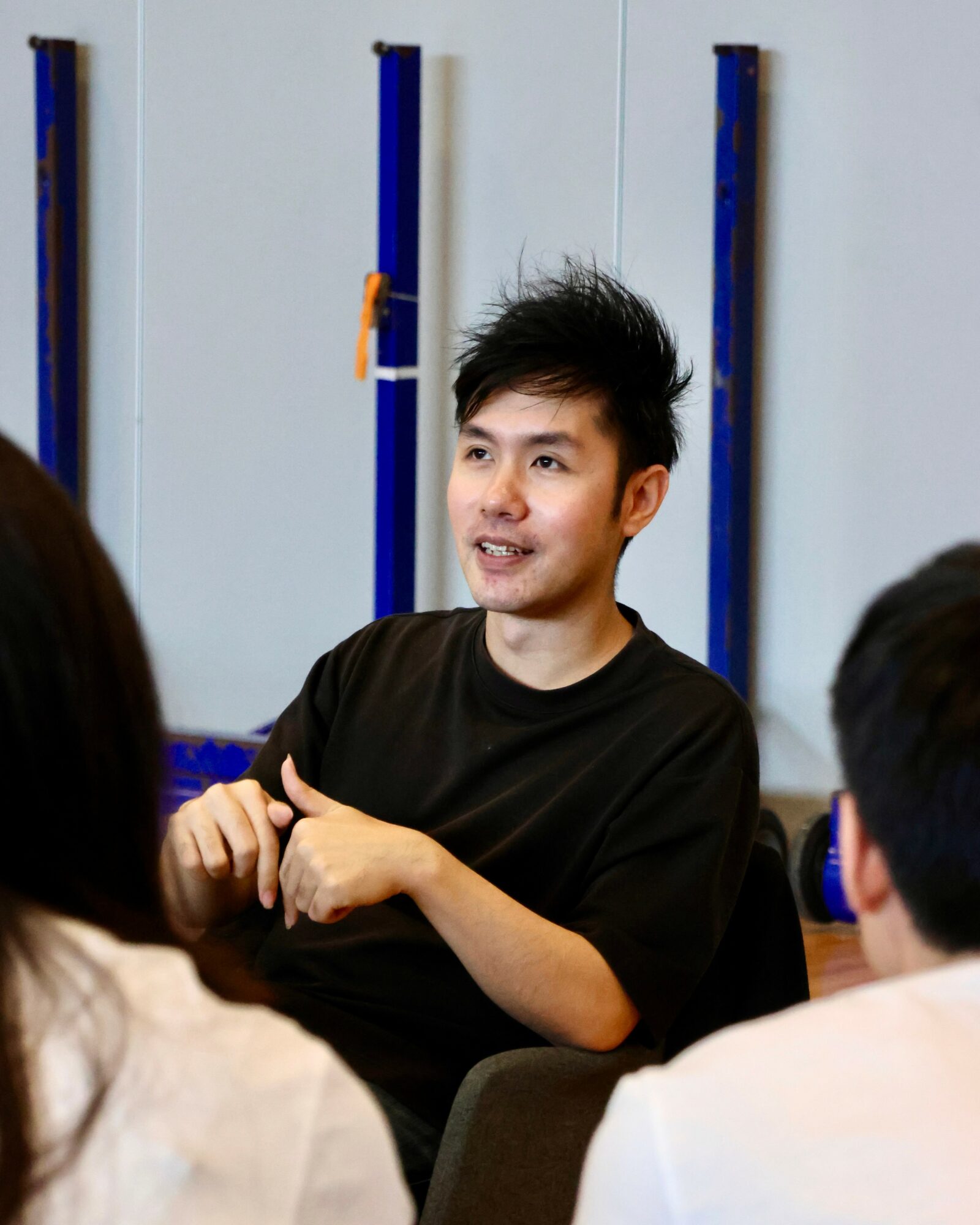 Young man with black hair speaking to a group, gesturing with hands, in a room with blue vertical bars on the wall.