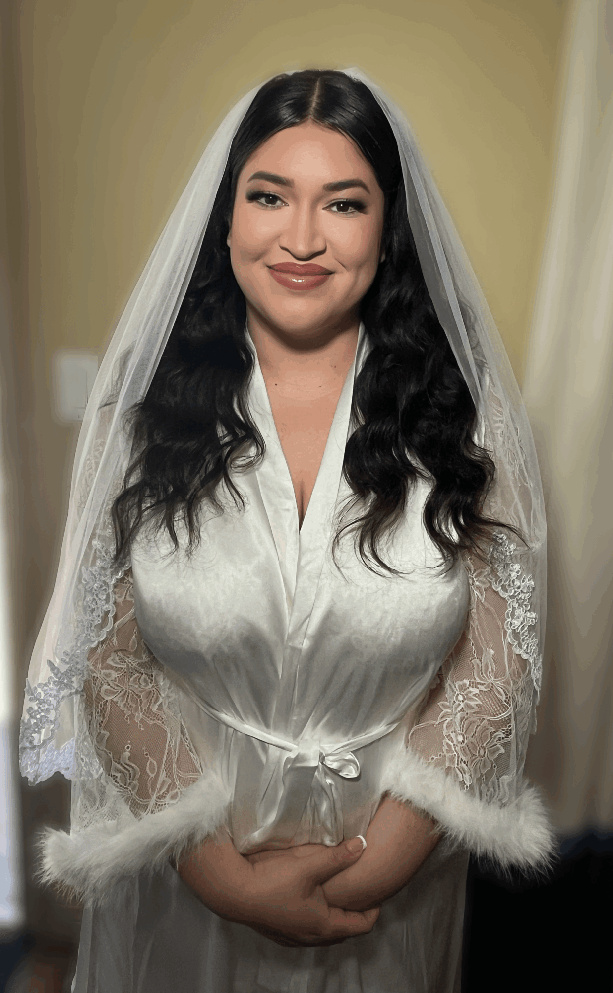 Woman in wedding dress with veil, smiling, dark wavy hair, standing indoors against a neutral background.