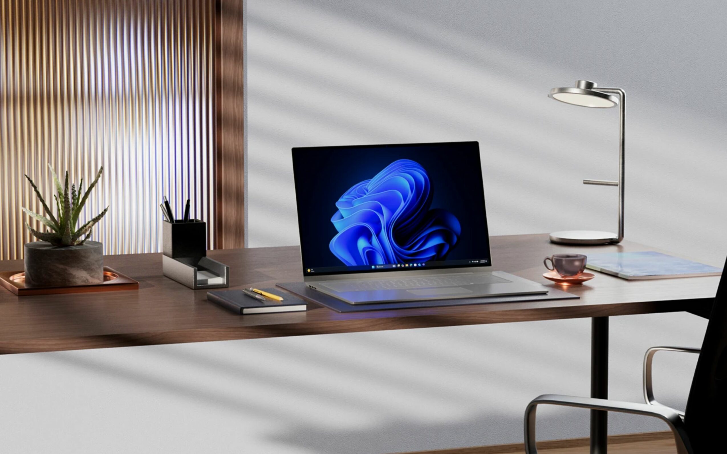 Office desk with a laptop, desk lamp, potted plant, and notebooks, illuminated by sunlight with shadows on the wall.