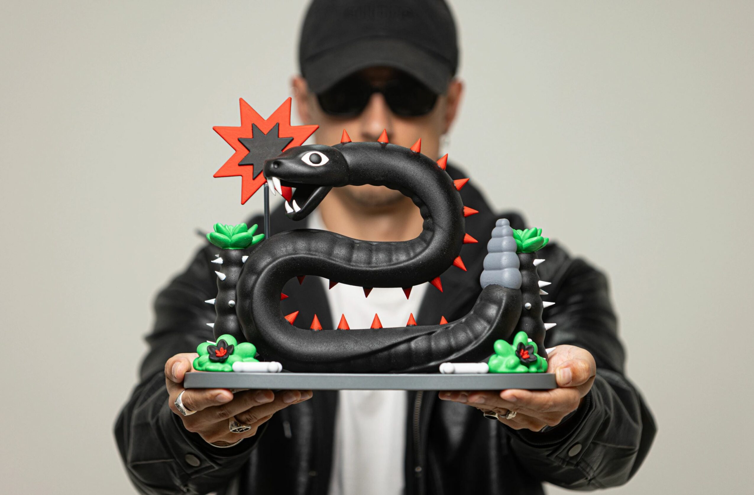 Person wearing sunglasses and a cap holding a black dragon sculpture with red spikes and green accents, against a plain background.