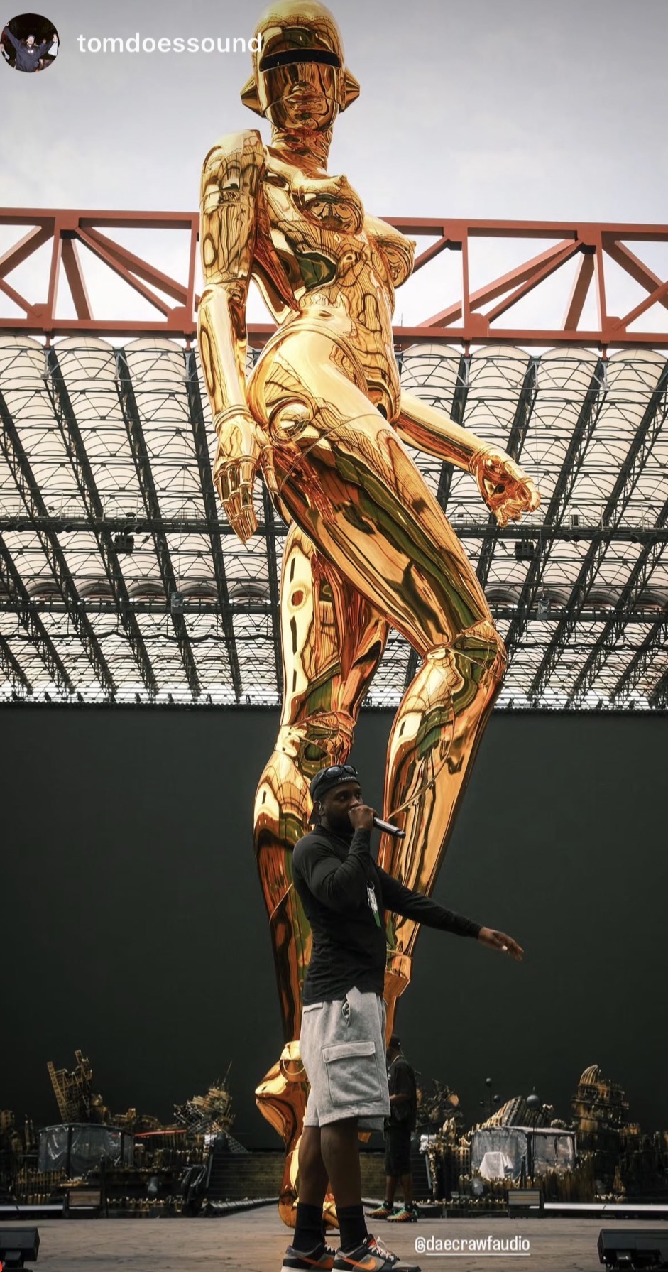 A person in a black shirt and white shorts stands in front of a large golden statue of a woman, with a bridge in the background.