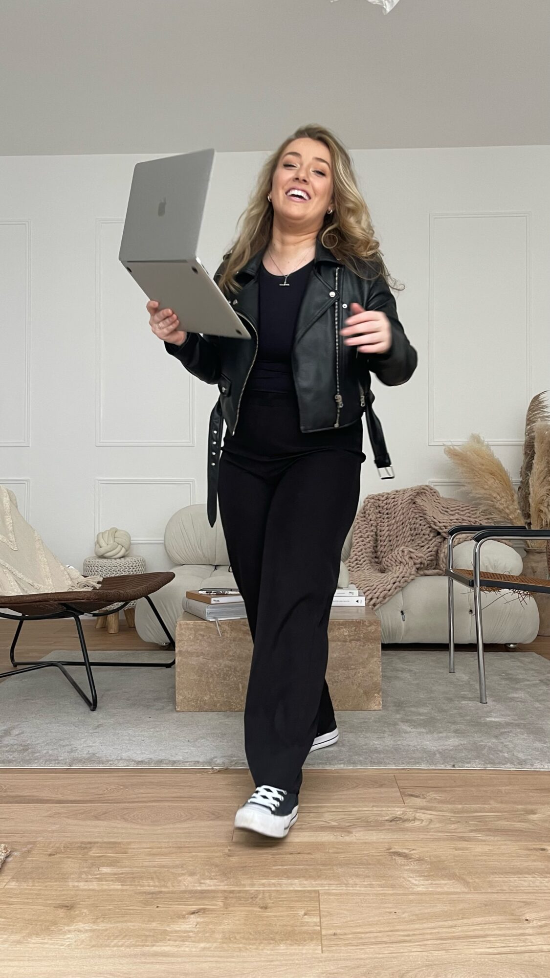 Woman walking indoors holding a laptop, smiling, in a living room with furniture and decor.