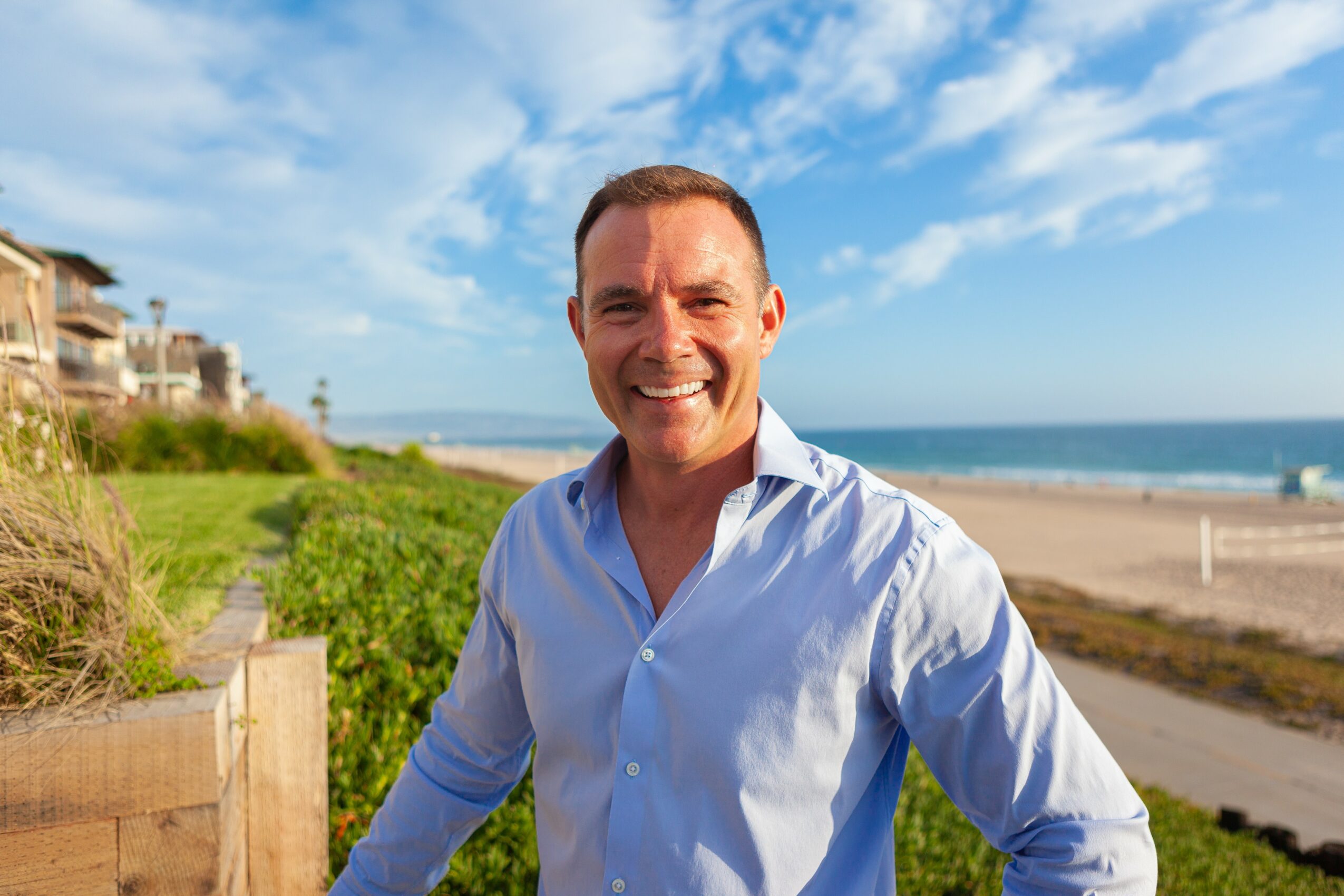 Smiling man in a light blue shirt standing outdoors near the beach with greenery and buildings in the background.