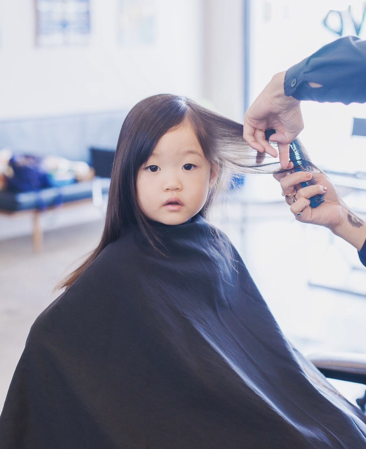 Child getting a haircut, sitting with a cape, hairdresser's hands cutting hair, bright salon interior in background.
