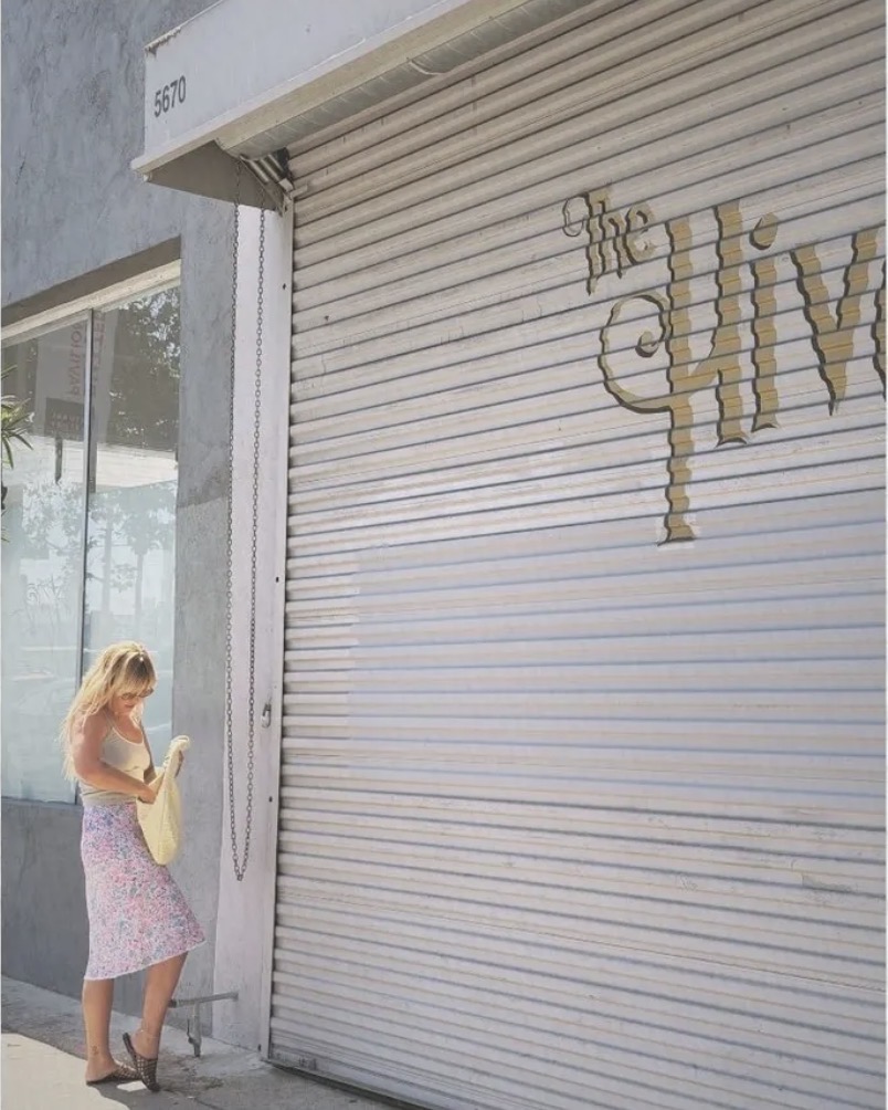 Woman with blonde hair in a pink dress standing outside a closed shop with a white metal shutter, holding a phone.