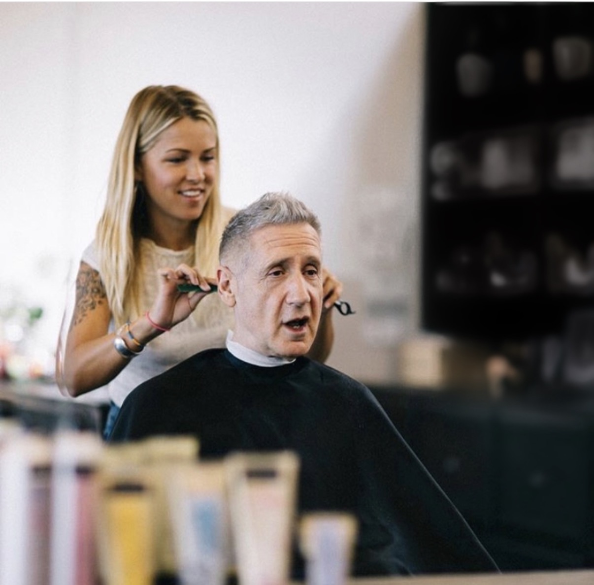 Young woman cutting an older man's hair in a salon, with shelves in the background.