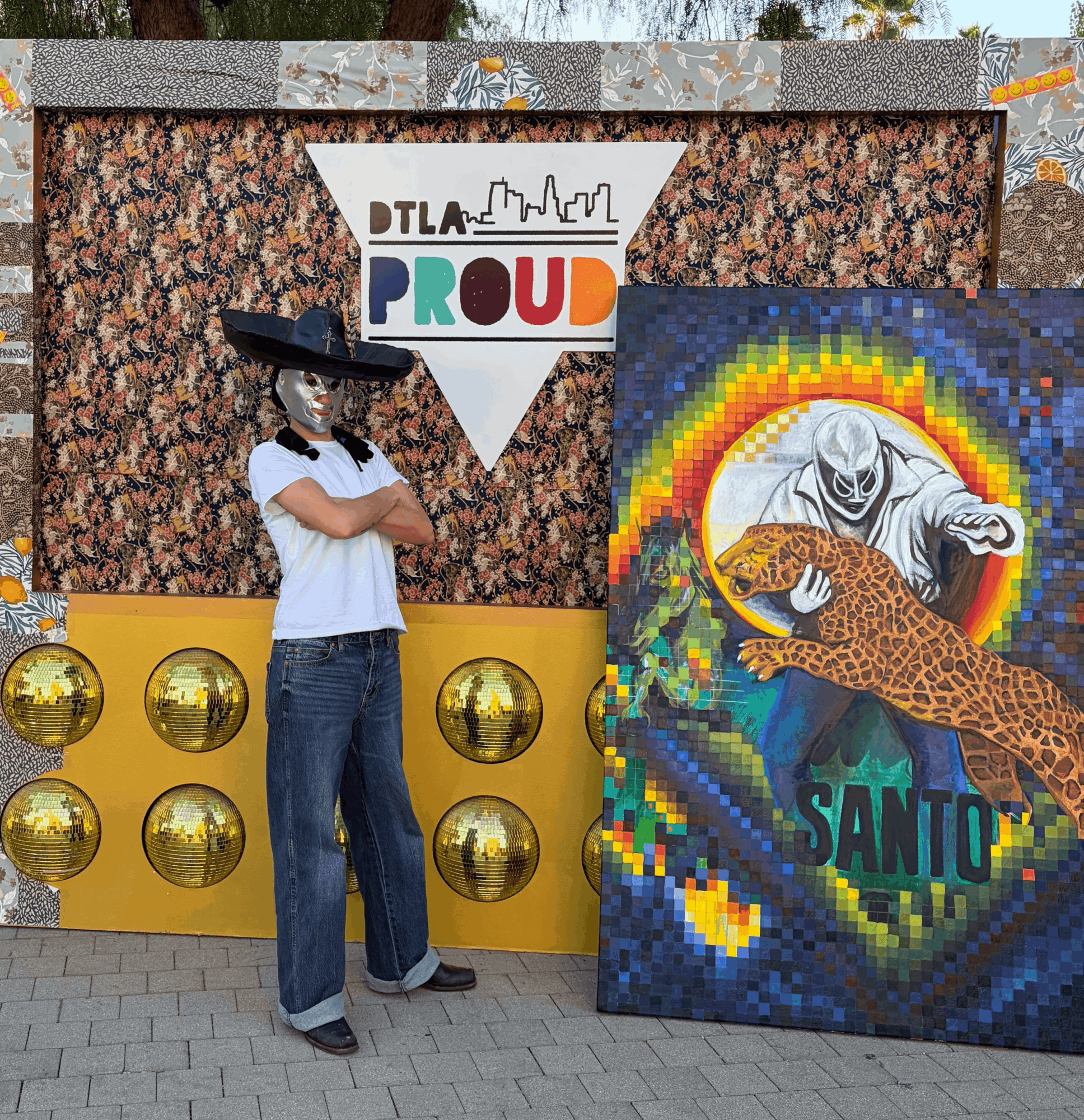 Person wearing a hat and white shirt standing with arms crossed next to colorful mosaic art and a sign that says 'Proud'.