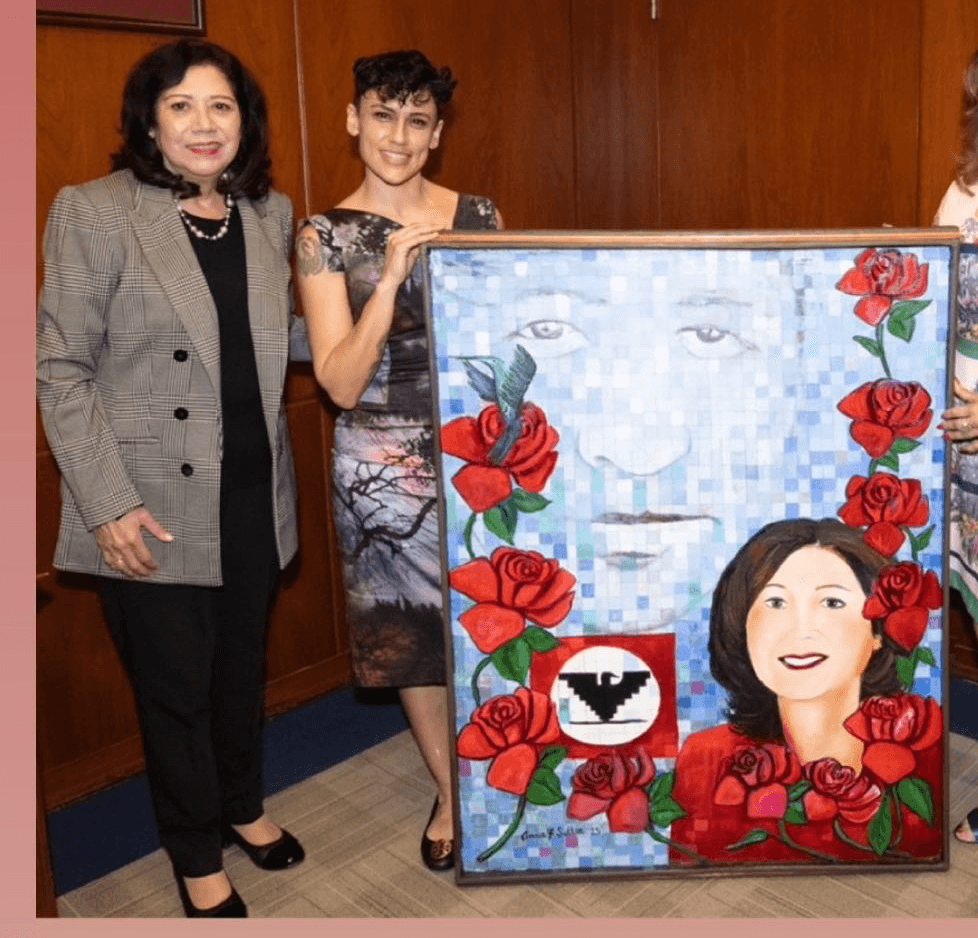 Two women stand next to a colorful portrait painting with a woman's face and red roses, indoors. Word 'VOTE' is visible.