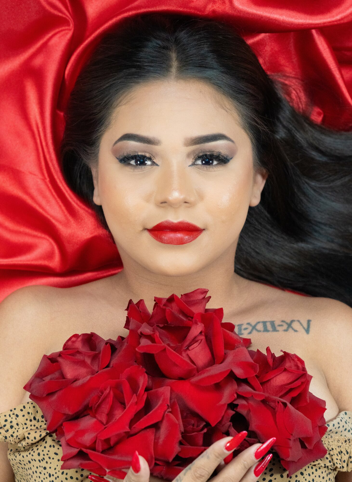 Woman with dark hair, makeup, red lipstick, lying on red satin fabric, holding red roses, with tattoo on shoulder.