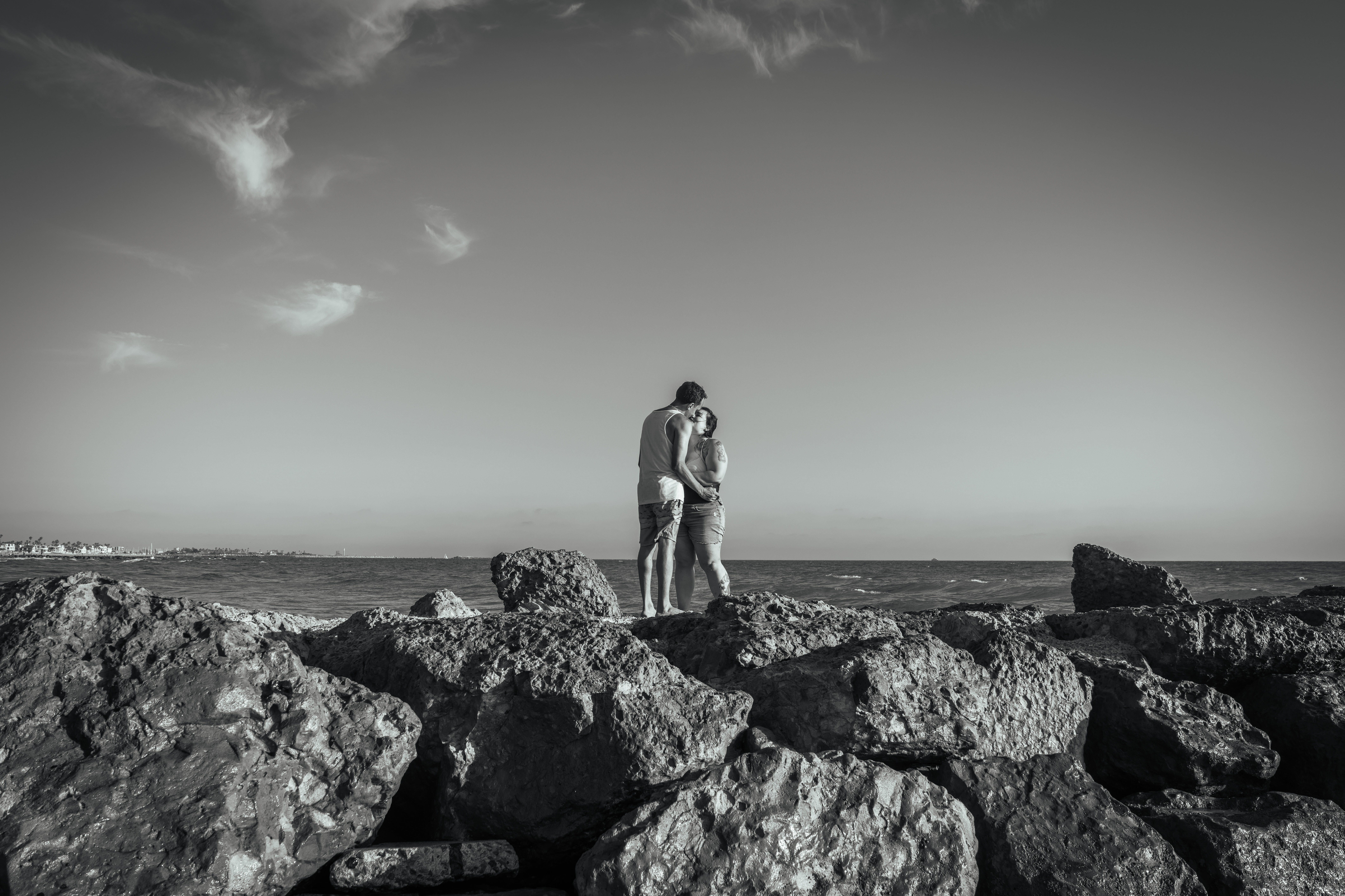 Two people standing on rocks near the ocean, with a cloudy sky in the background.