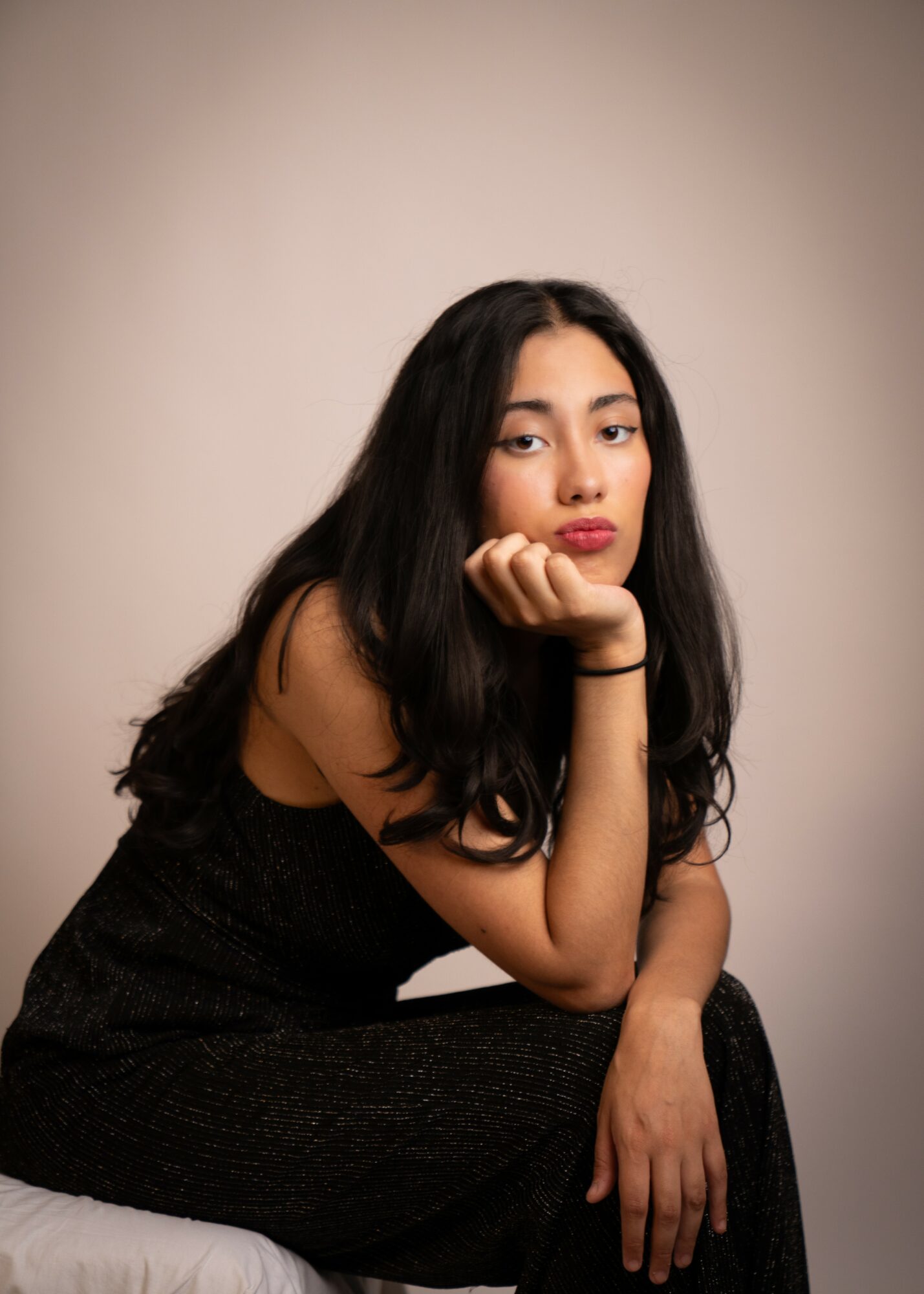 Young woman with long dark hair resting her chin on her hand, wearing a black dress, against a plain background.