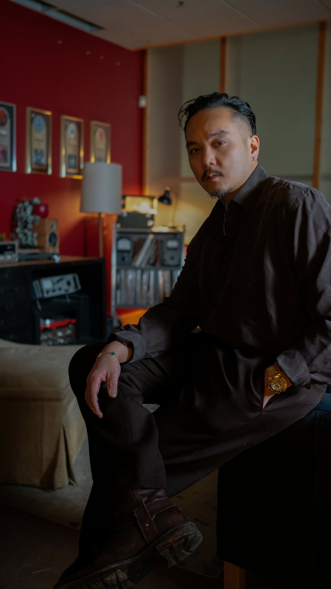 Man sitting on a dark piece of furniture in a room with red and green walls, books, and framed pictures, looking at the camera.