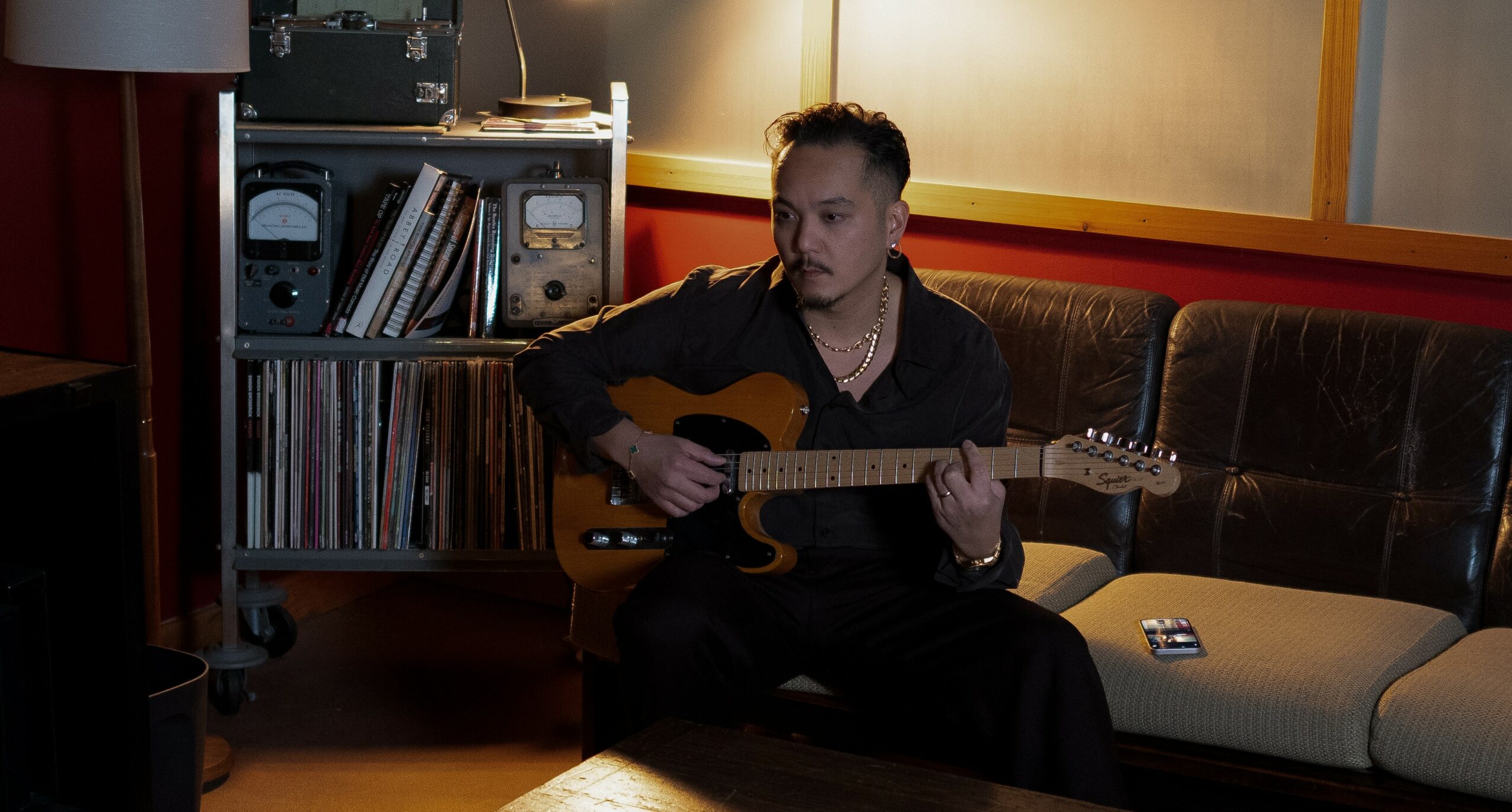 Person sitting on a sofa playing an acoustic guitar in a room with books and records.
