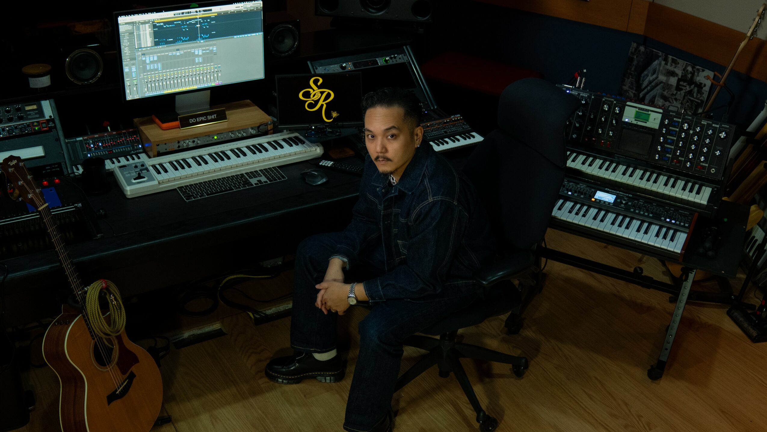 Young person sitting in a music studio surrounded by keyboards, computers, and a guitar, looking at the camera.
