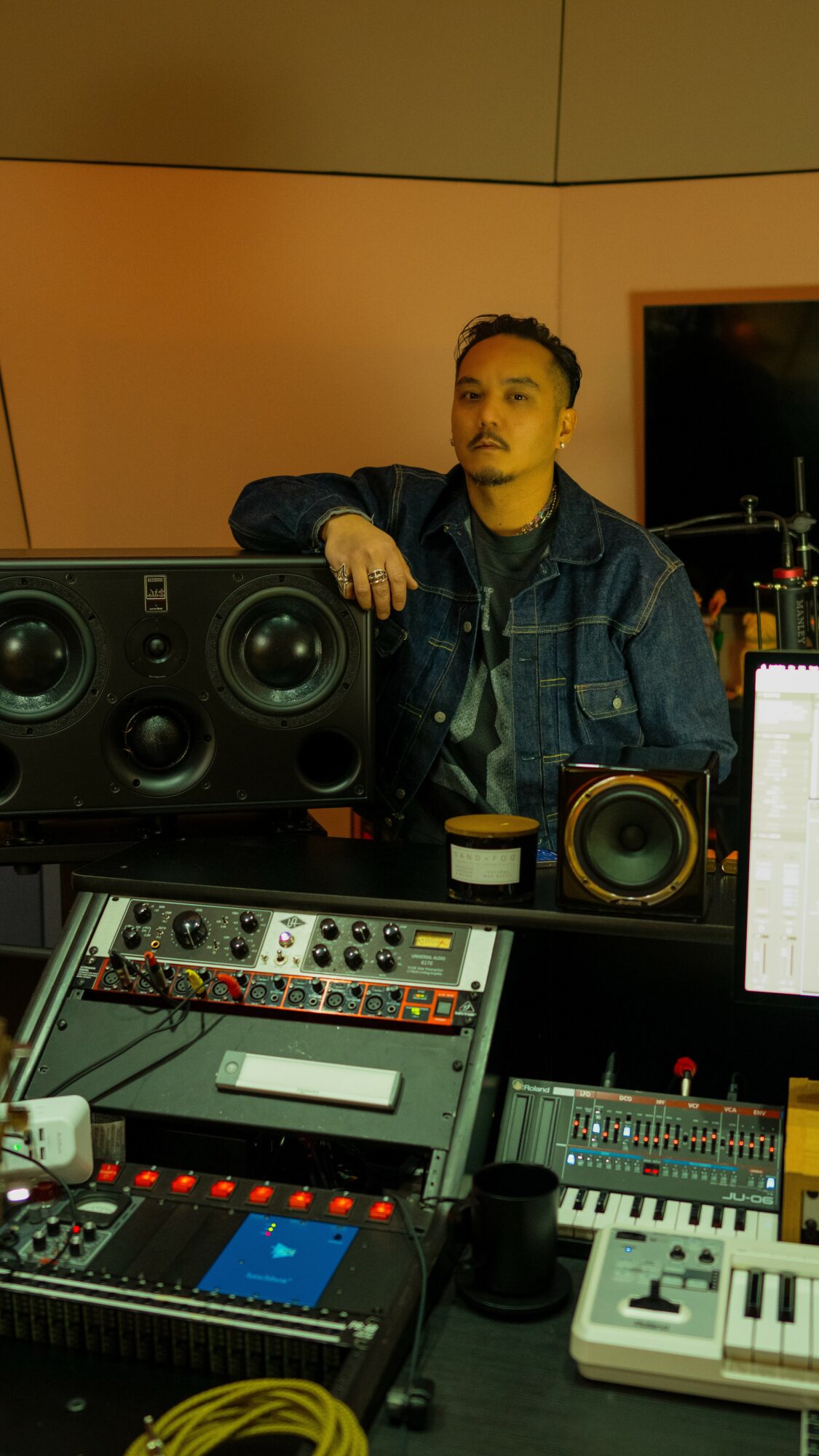 Young man with short hair and a beard stands behind audio equipment in a recording studio, wearing a denim jacket.