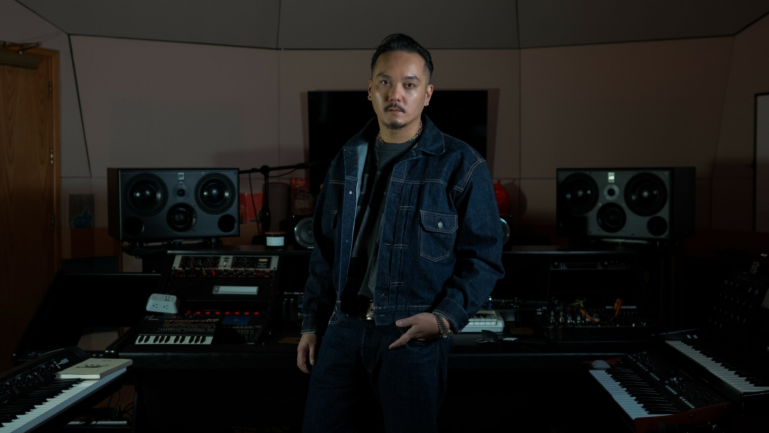 Person standing in a music studio with audio equipment and keyboards, wearing a denim jacket and looking at the camera.
