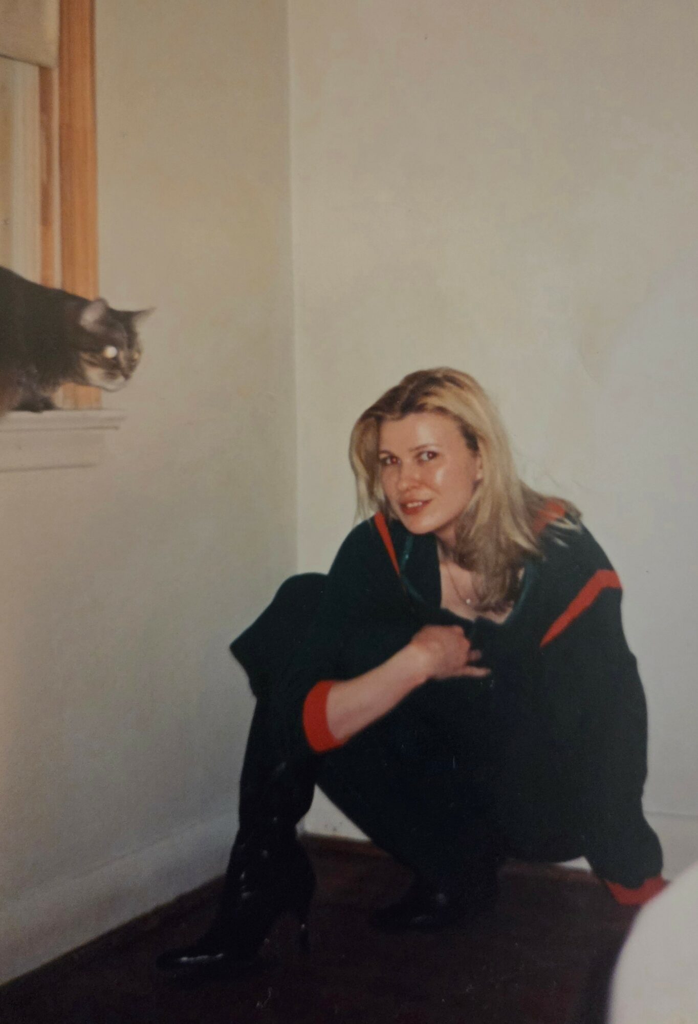 Woman crouching near a wall with a cat on a window ledge, smiling at the camera.
