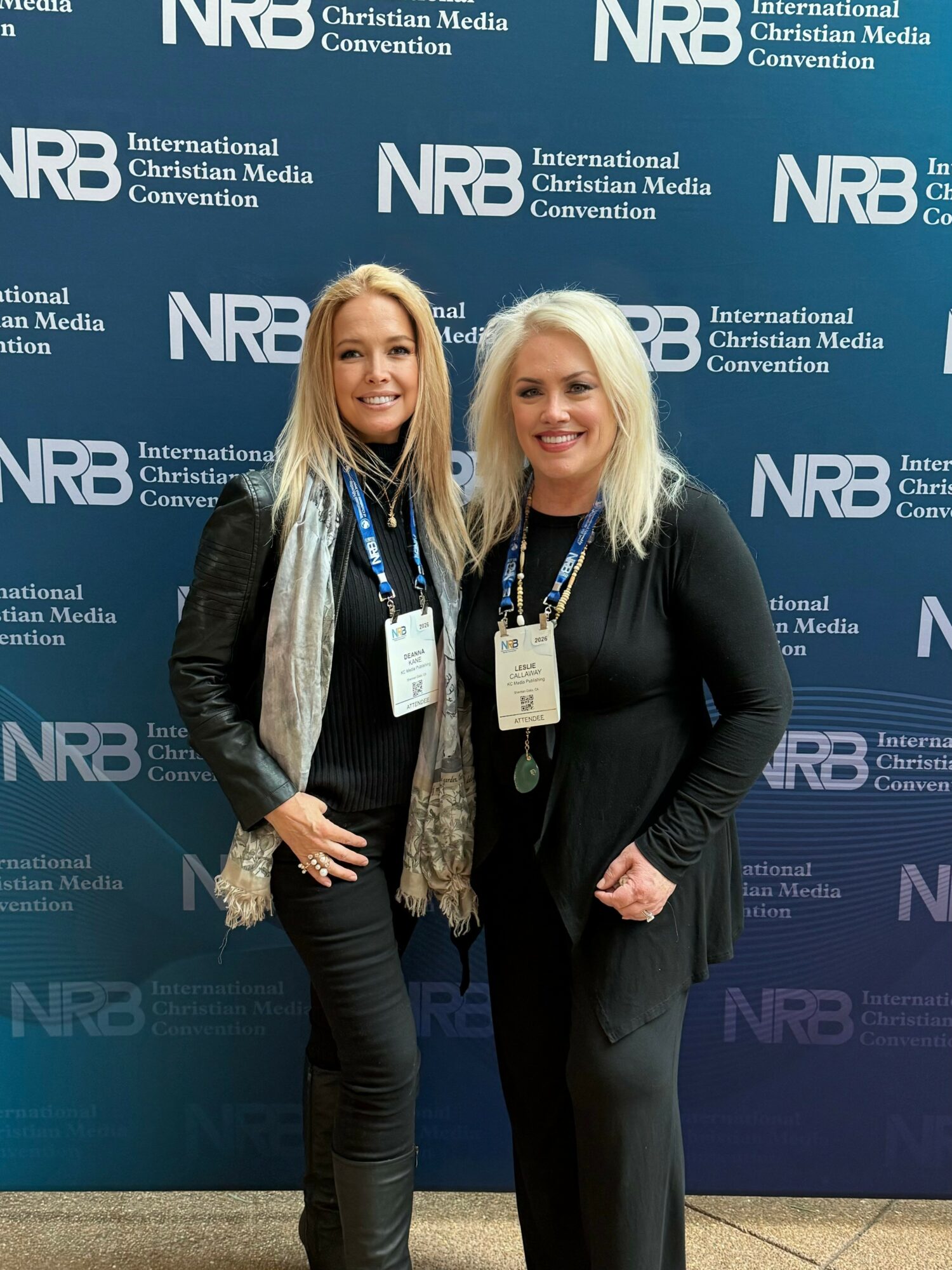 Two women standing together in front of a blue backdrop with white text, smiling, wearing conference badges.