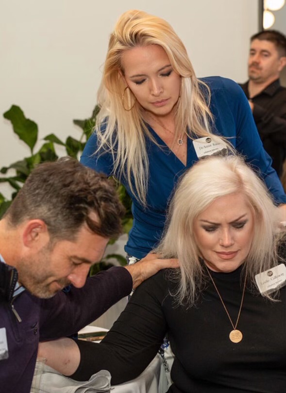 Three people in a room, woman with blonde hair leaning over, woman with white hair sitting, man with dark hair and beard sitting, plants in background.