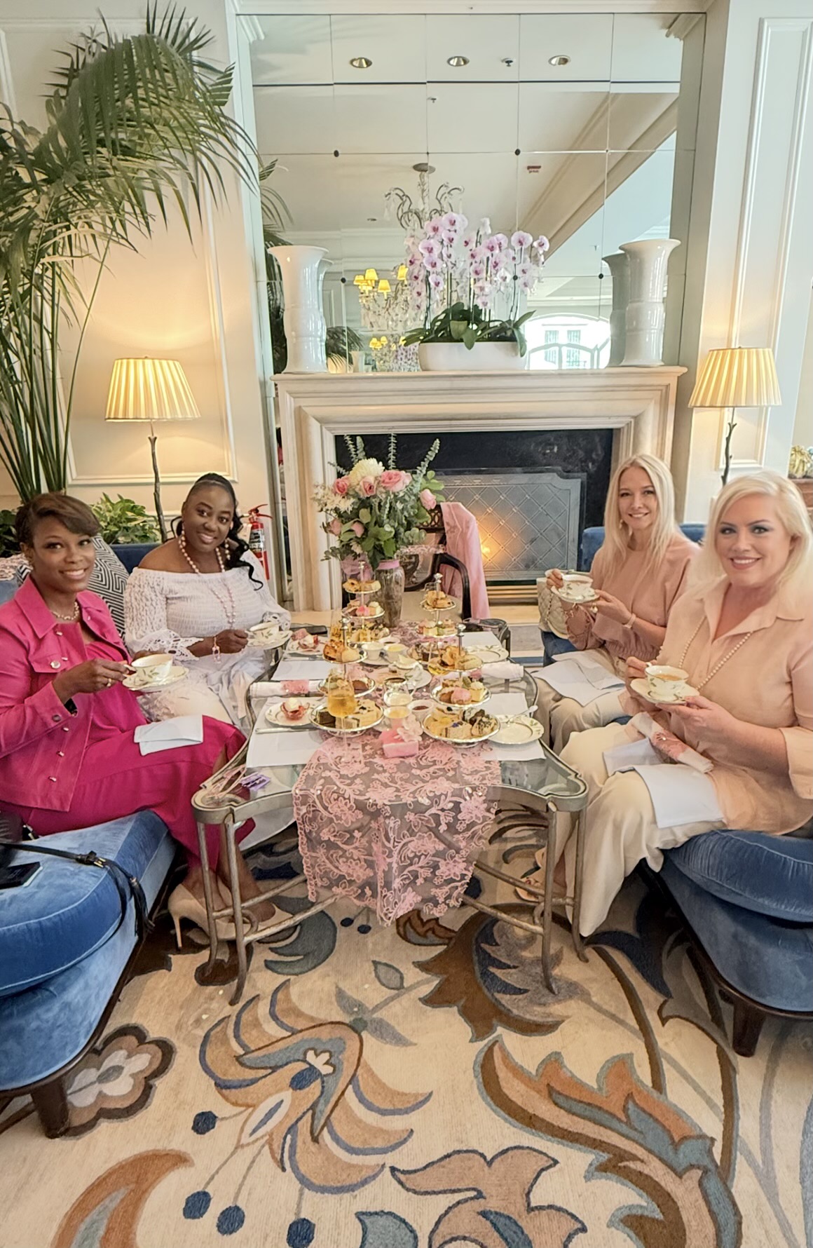 Four women sit at a dining table with food and drinks, in a bright room with a fireplace and floral decor.