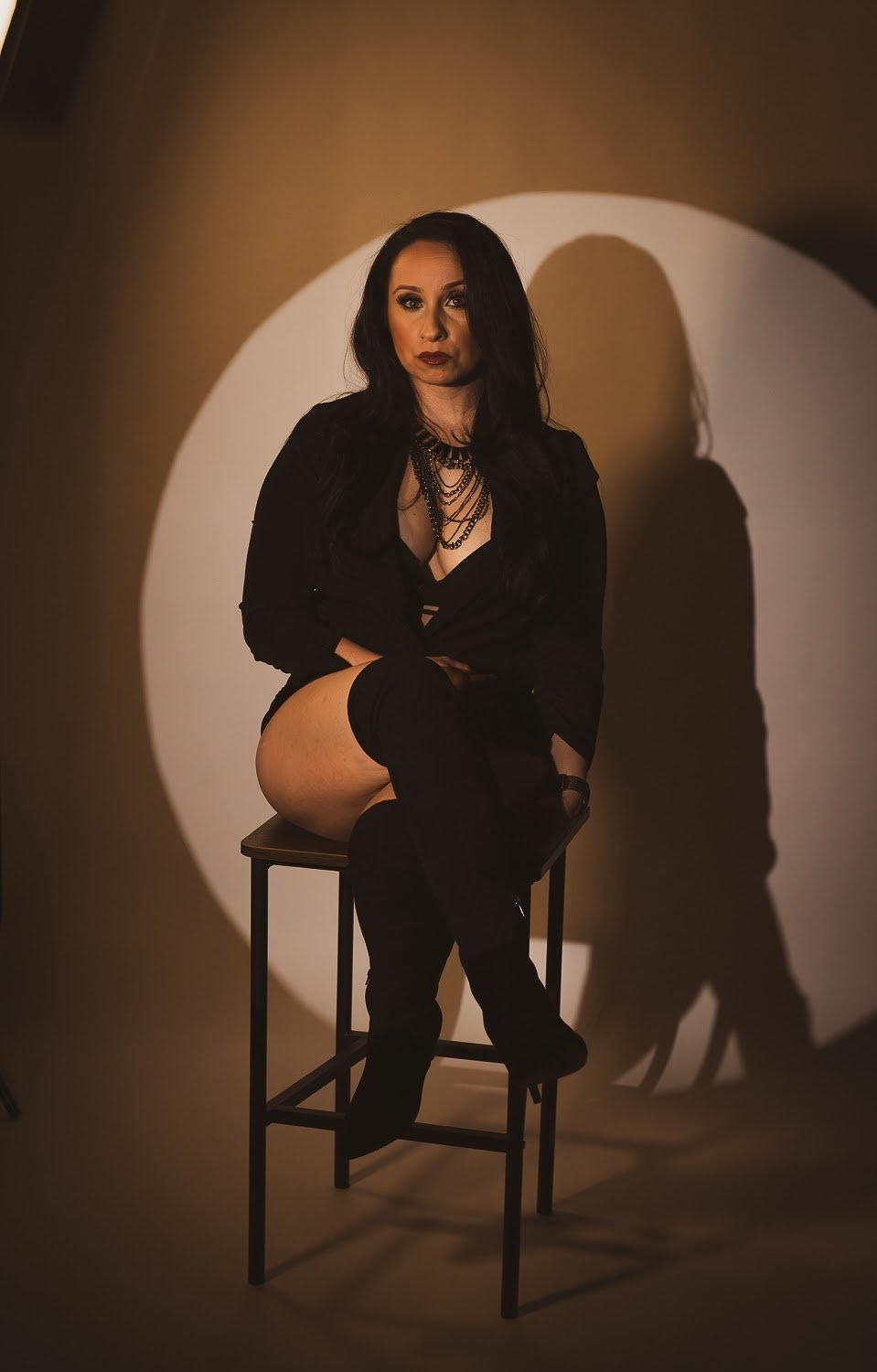 Woman sitting on a stool with a shadow cast on the wall behind her, wearing black clothing and thigh-high boots.