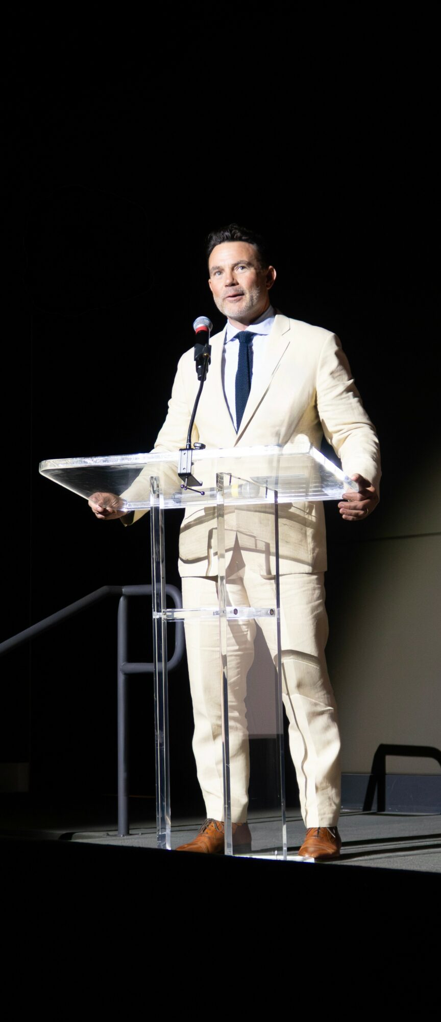 Man in a white suit and blue tie speaking at a clear podium on stage, with a dark background.