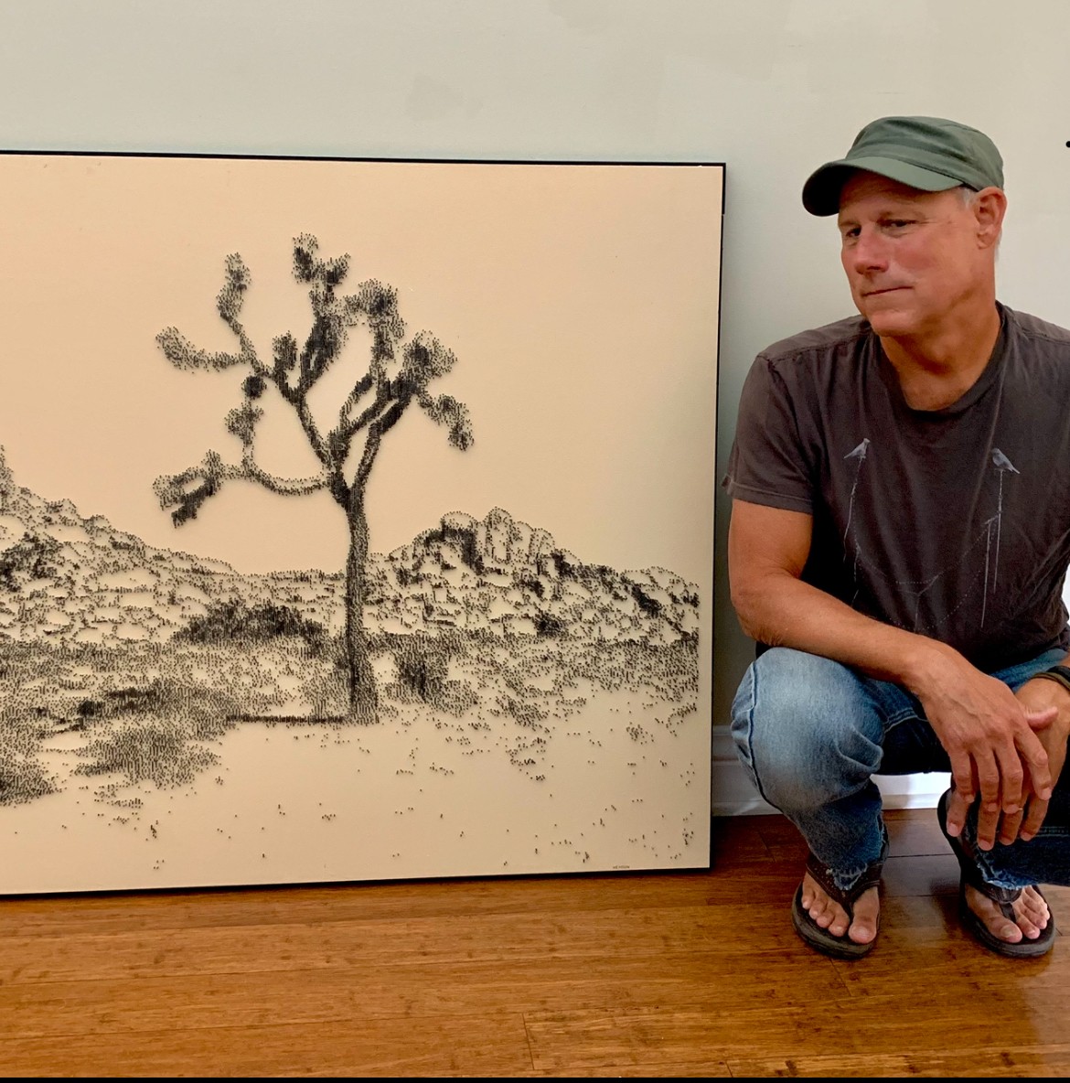Person sitting next to a framed black and white landscape painting of a tree and rocky terrain, on a wooden floor.