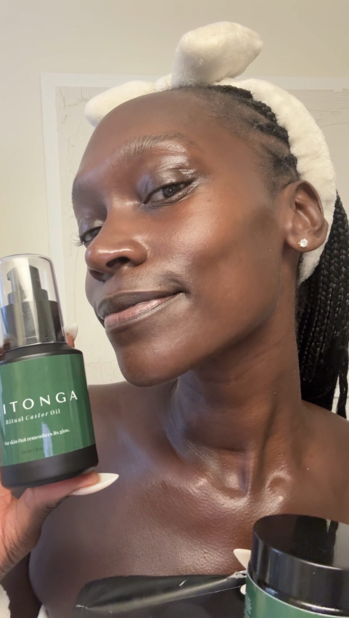 Woman with braided hair and a headband holding a green skincare product bottle, smiling slightly.