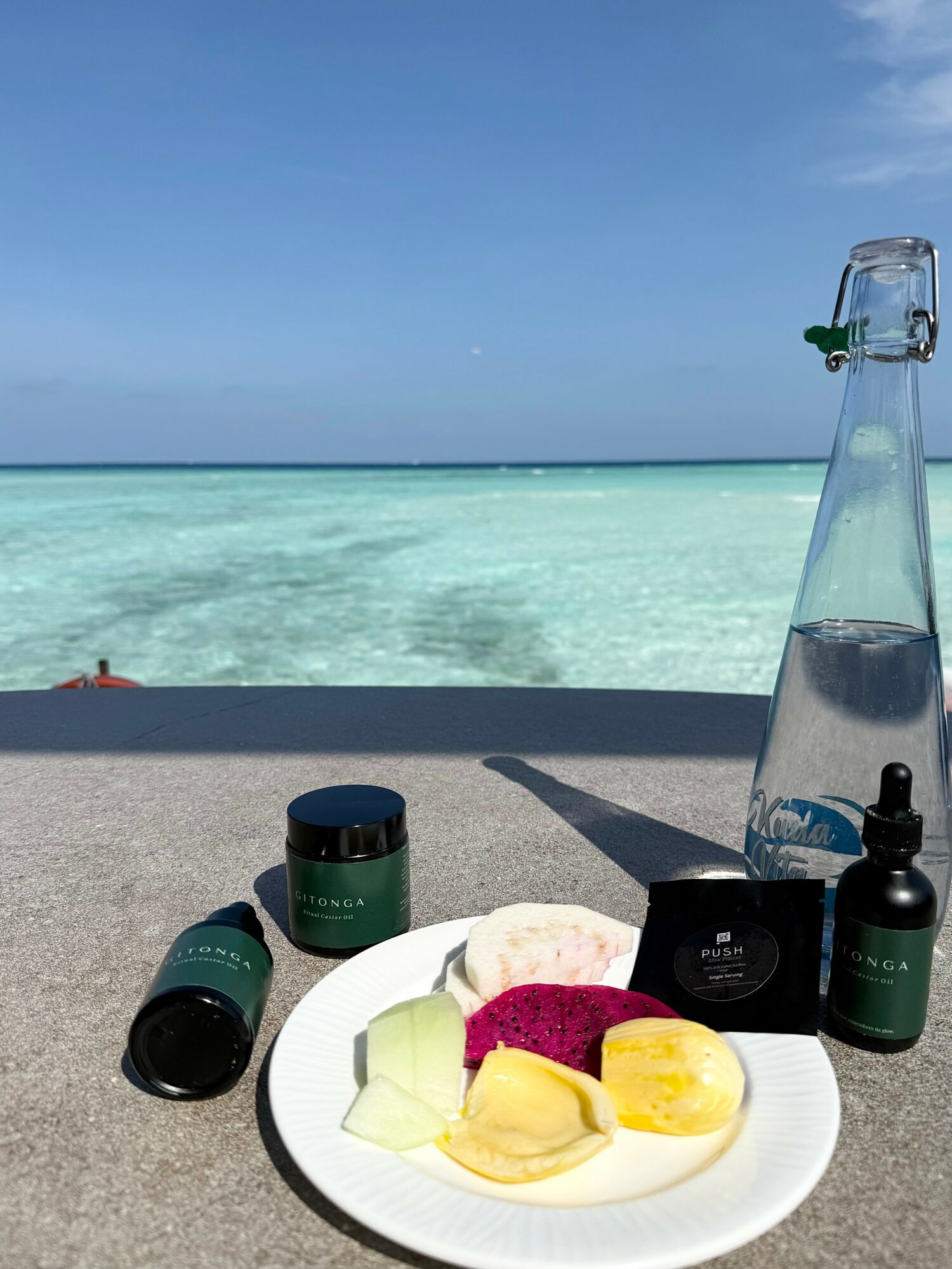 Beach scene with clear water, blue sky, and a table with skincare products and a plate of fruit, including banana and dragon fruit.