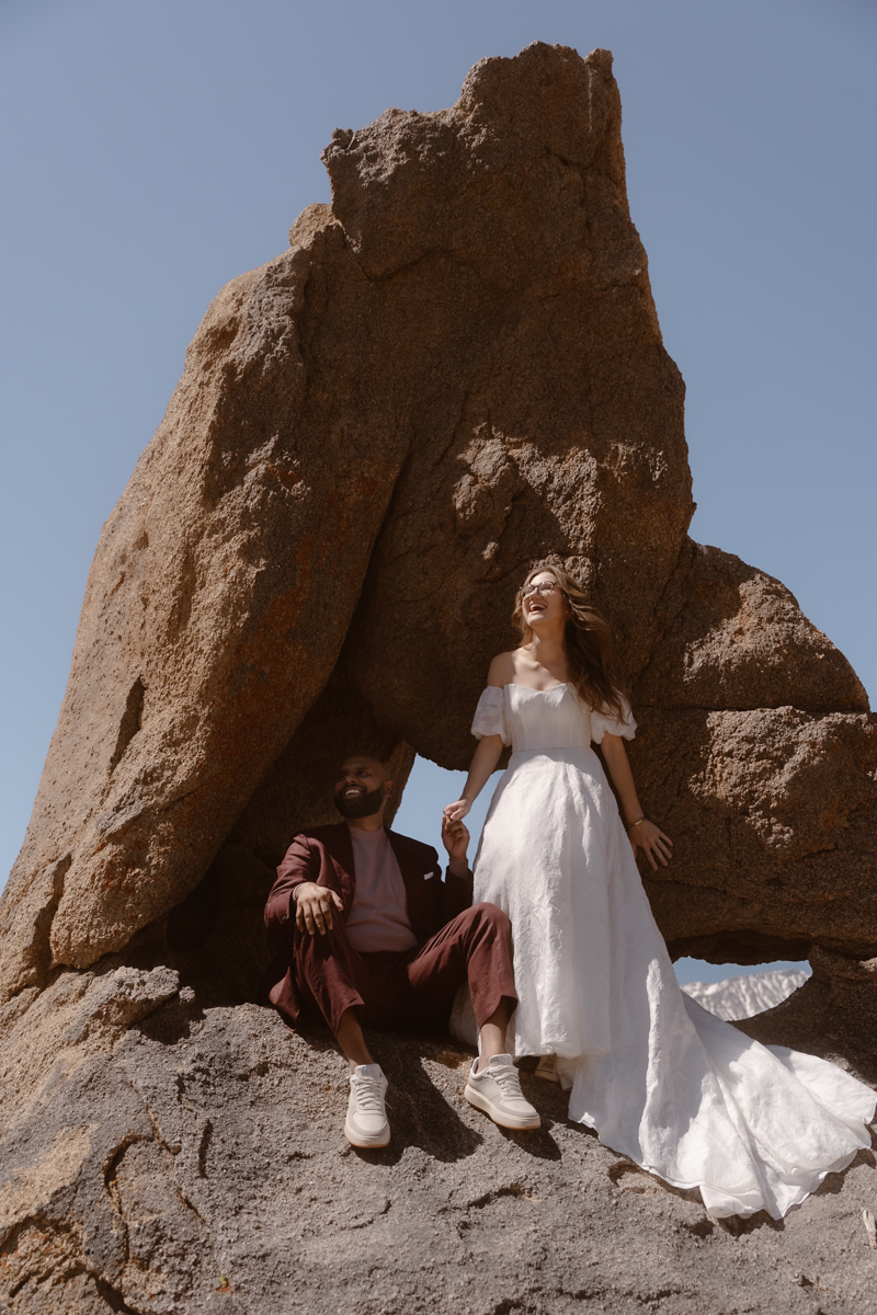 A woman in a white dress and a man in a suit sit on rocks near a large rock formation, holding hands.