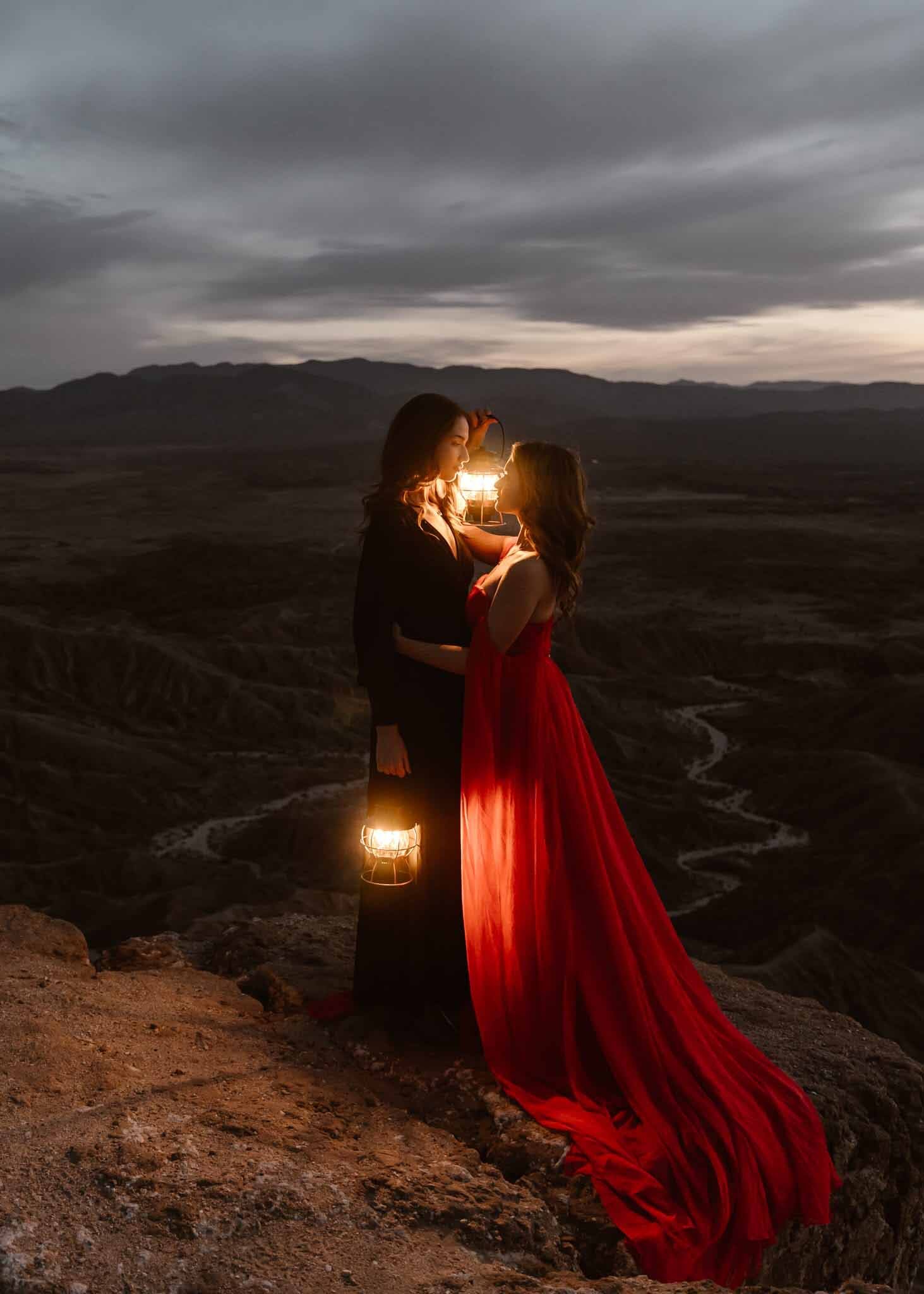 Two women in elegant dresses stand close, holding a lantern and a glowing object, with a vast landscape and cloudy sky behind them.