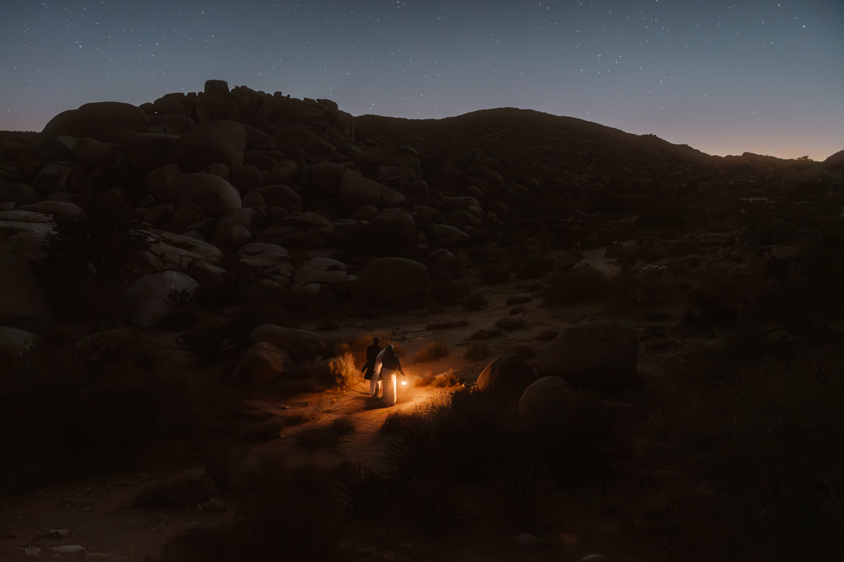 Two people stand near a small campfire in a dark, rocky landscape under a starry sky.