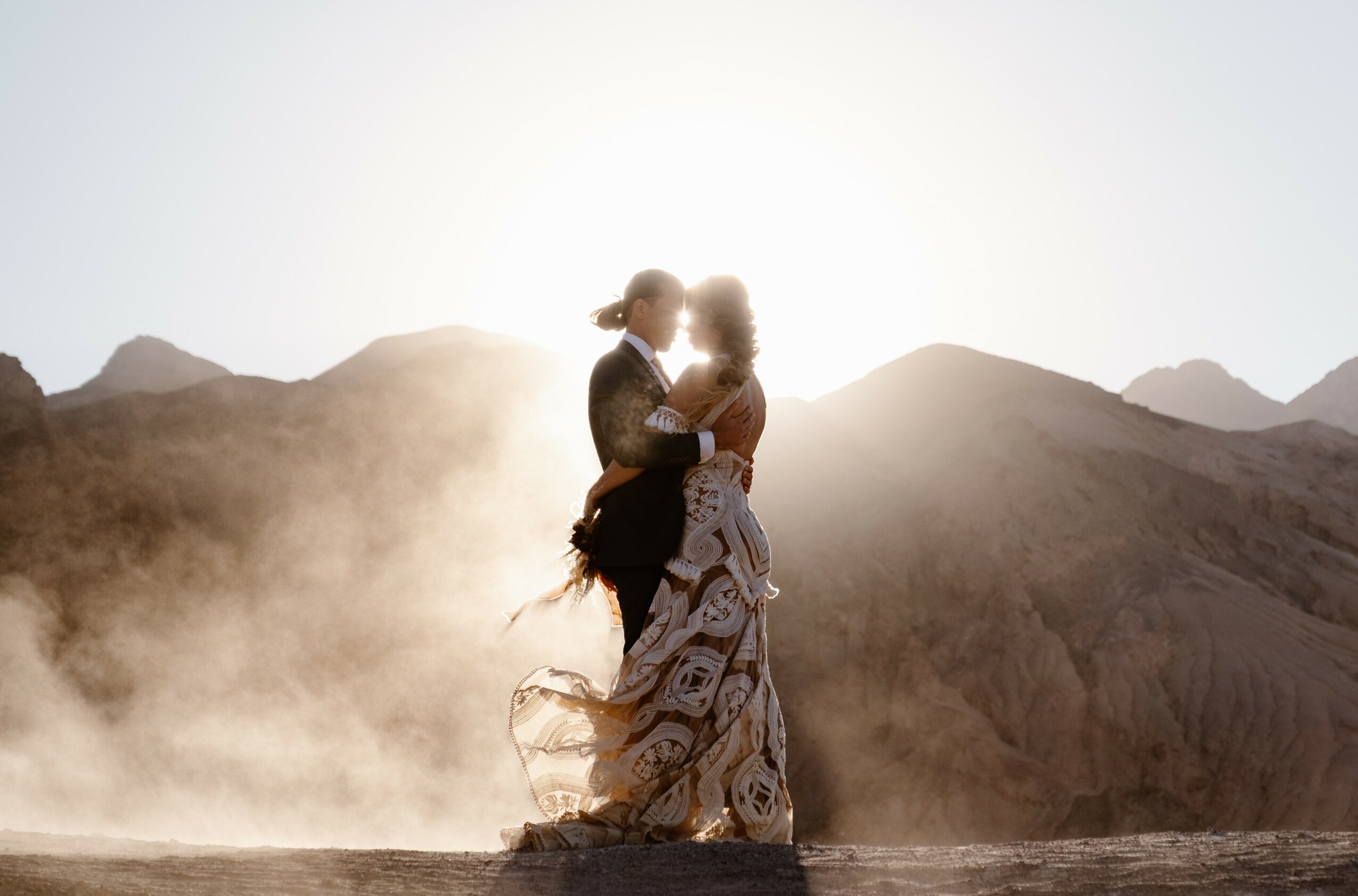 Two people embrace in a desert landscape with mountains and dust, backlit by the sun, creating a silhouette effect.