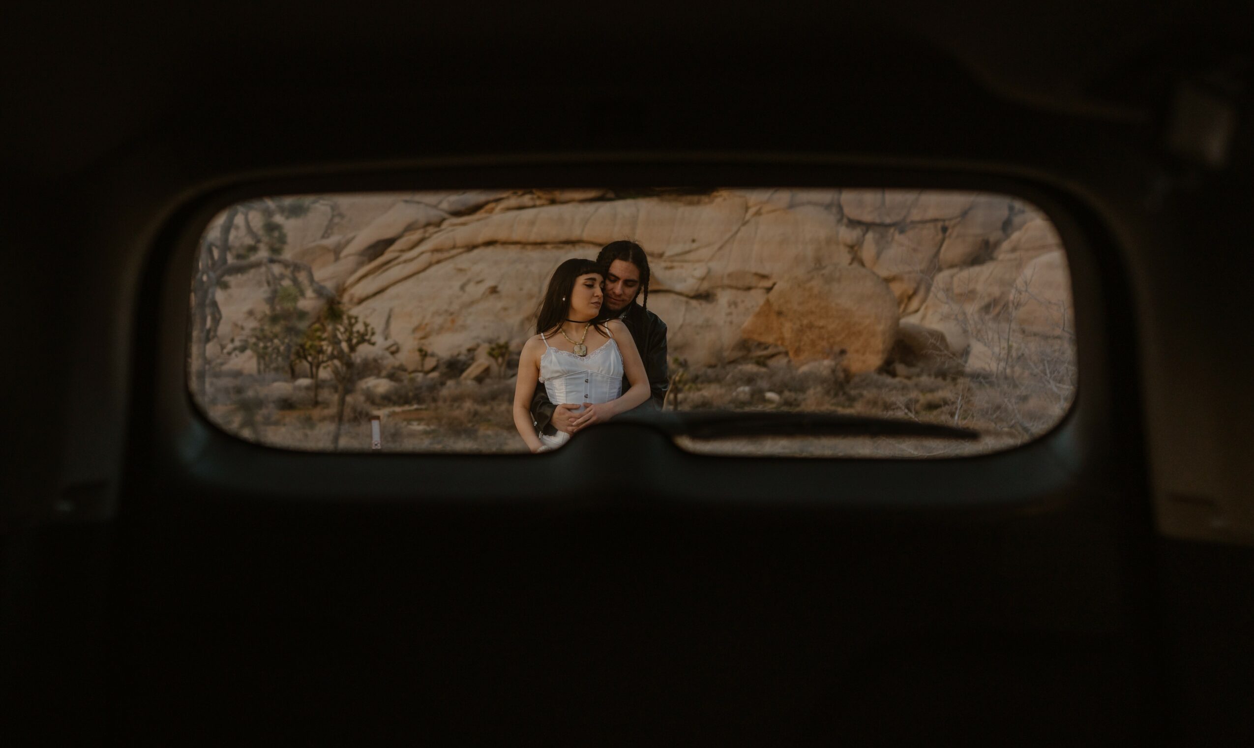 Two women standing outdoors near rocks, viewed through a car's rear window, with a natural landscape background.