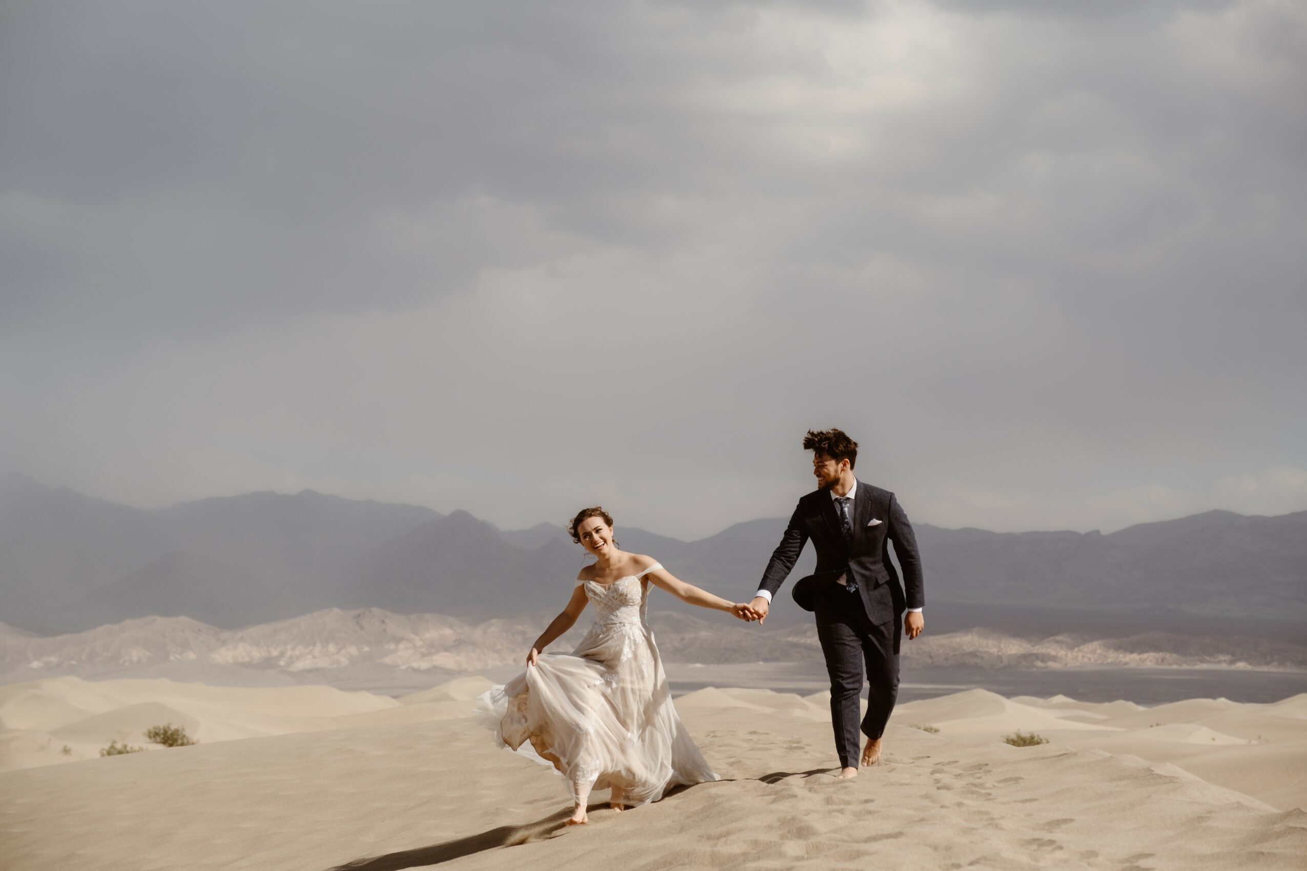A couple holding hands walking on a sandy landscape under cloudy sky, mountains in the background.