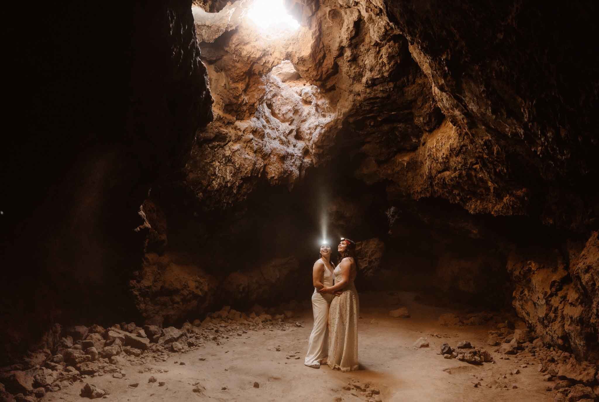 Two people stand inside a cave, holding a light, with rocky walls and a bright opening above.