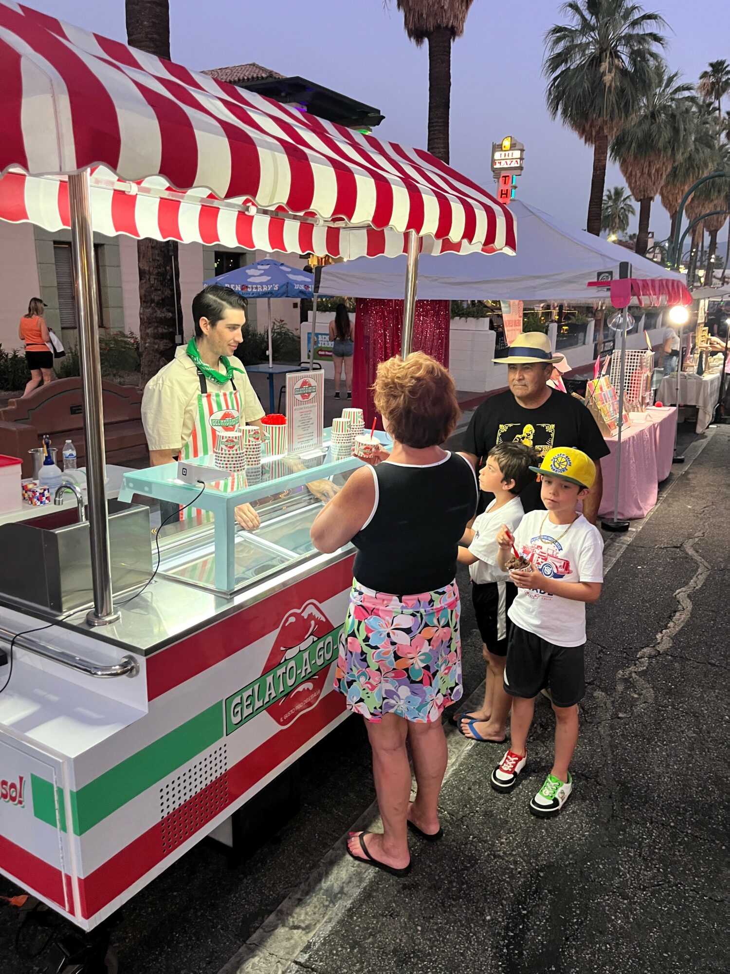 People at a food stand with a red and white striped canopy, including children and a woman, outdoors with palm trees.