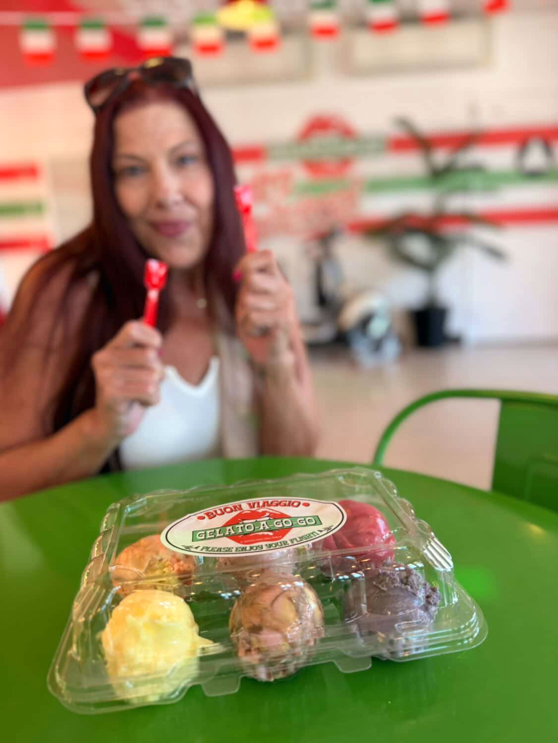 Woman holding a red lollipop, smiling, with a plastic container of assorted candies on a green table in front.