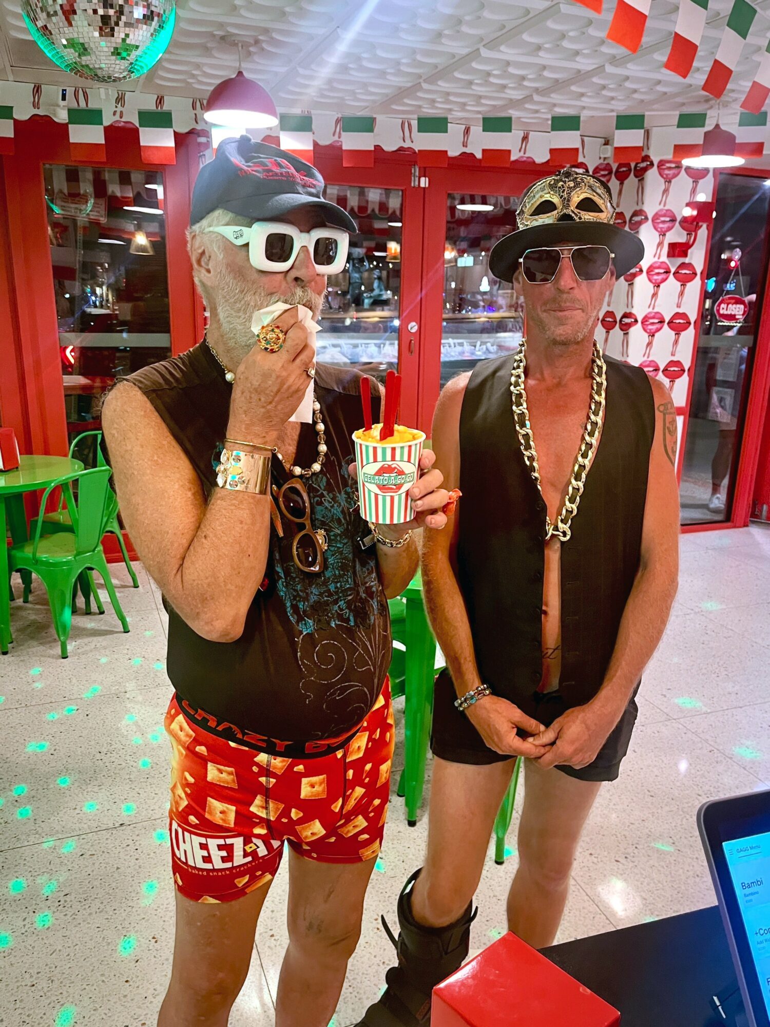 Two people in costumes with sunglasses, one eating ice cream, inside a colorful store with red and green decor.