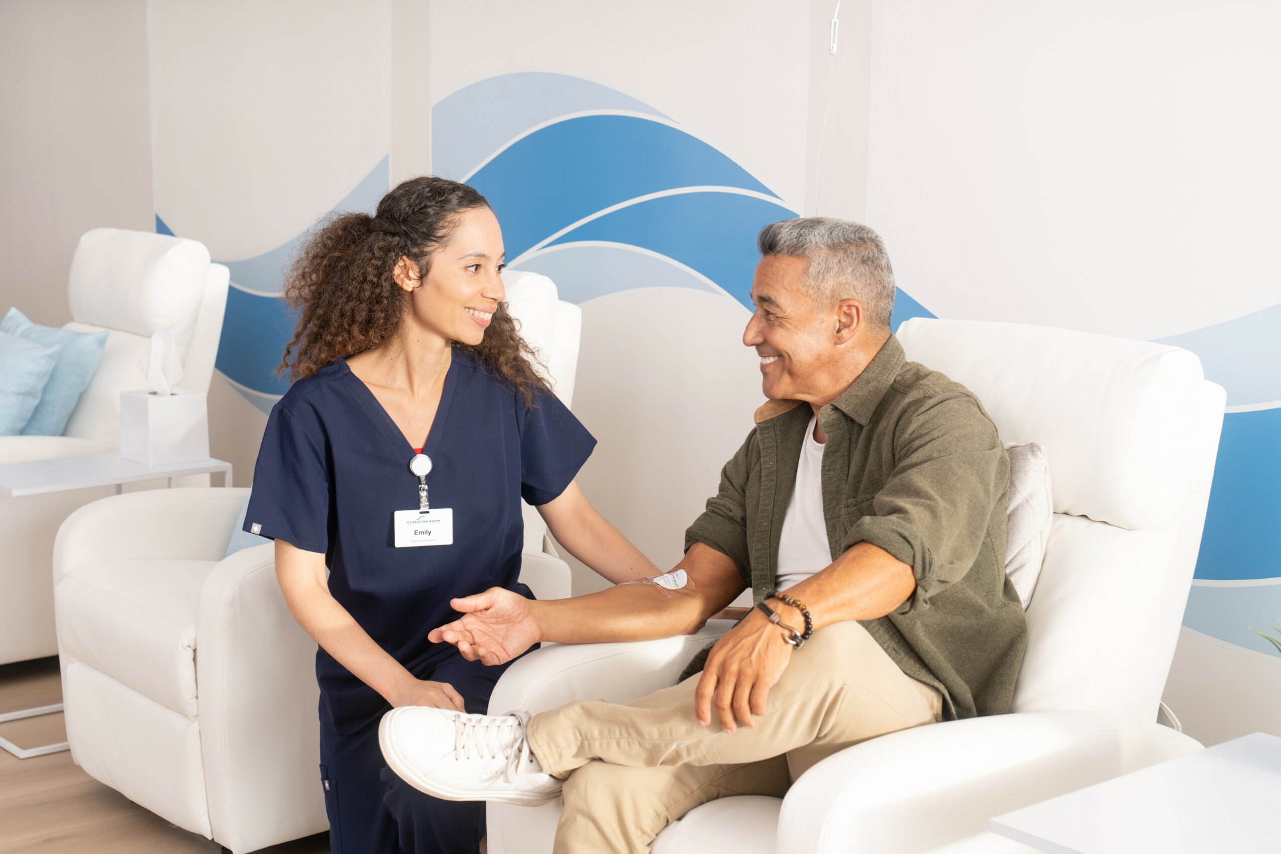 A nurse and a patient sit on white chairs, holding hands and smiling at each other in a medical setting.