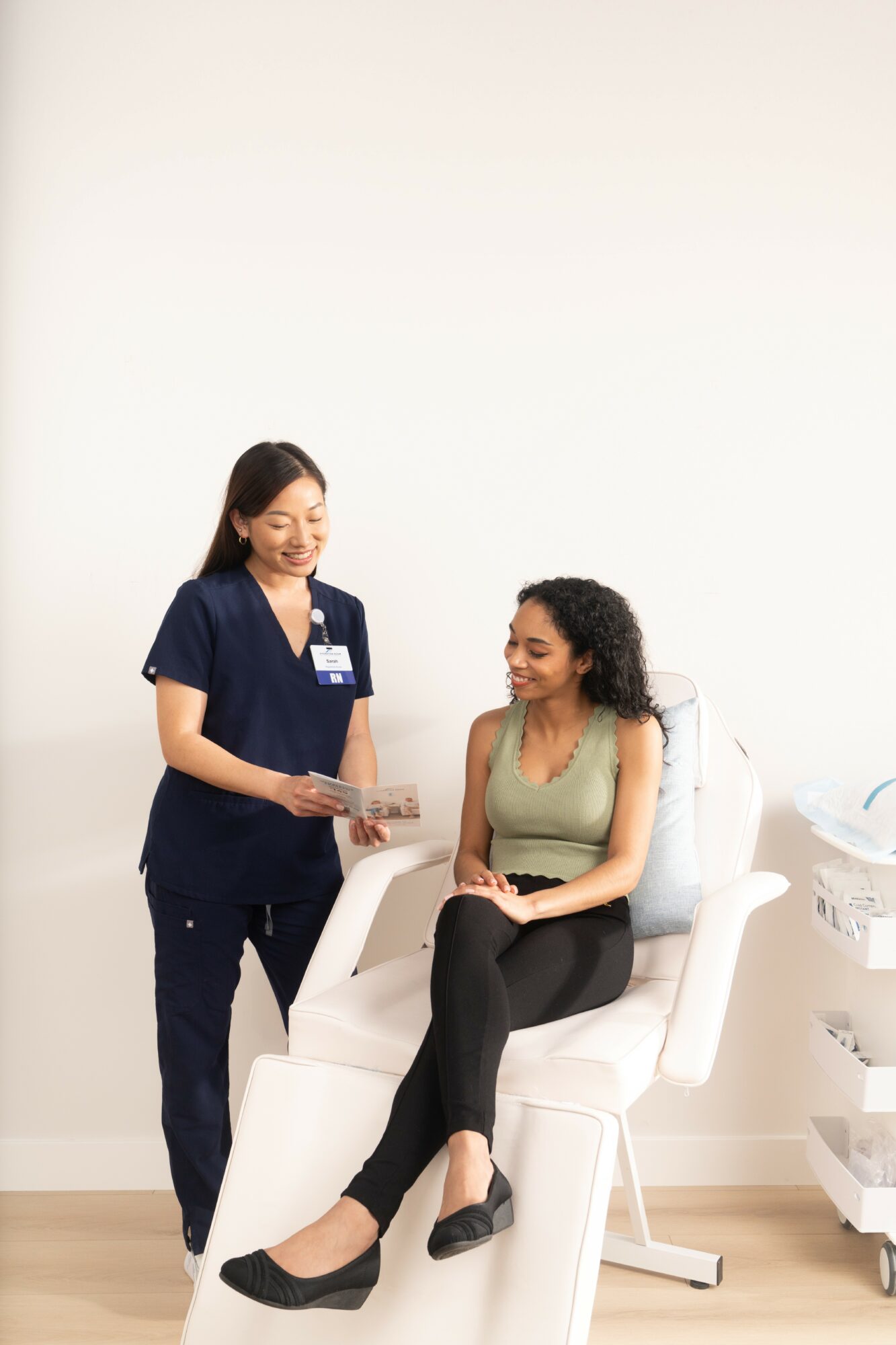 A woman in medical scrubs standing and talking to a seated woman in a medical setting, both smiling.