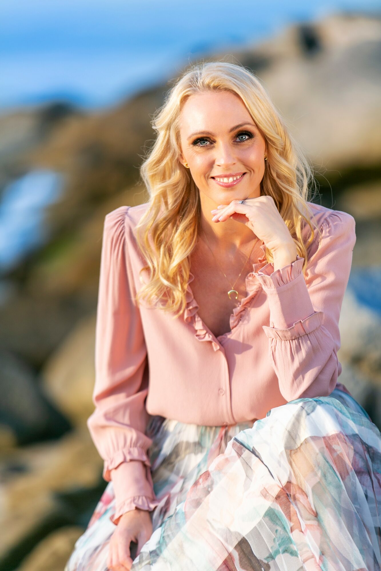 Smiling woman with blonde hair in a pink blouse and patterned skirt outdoors near rocks and water.