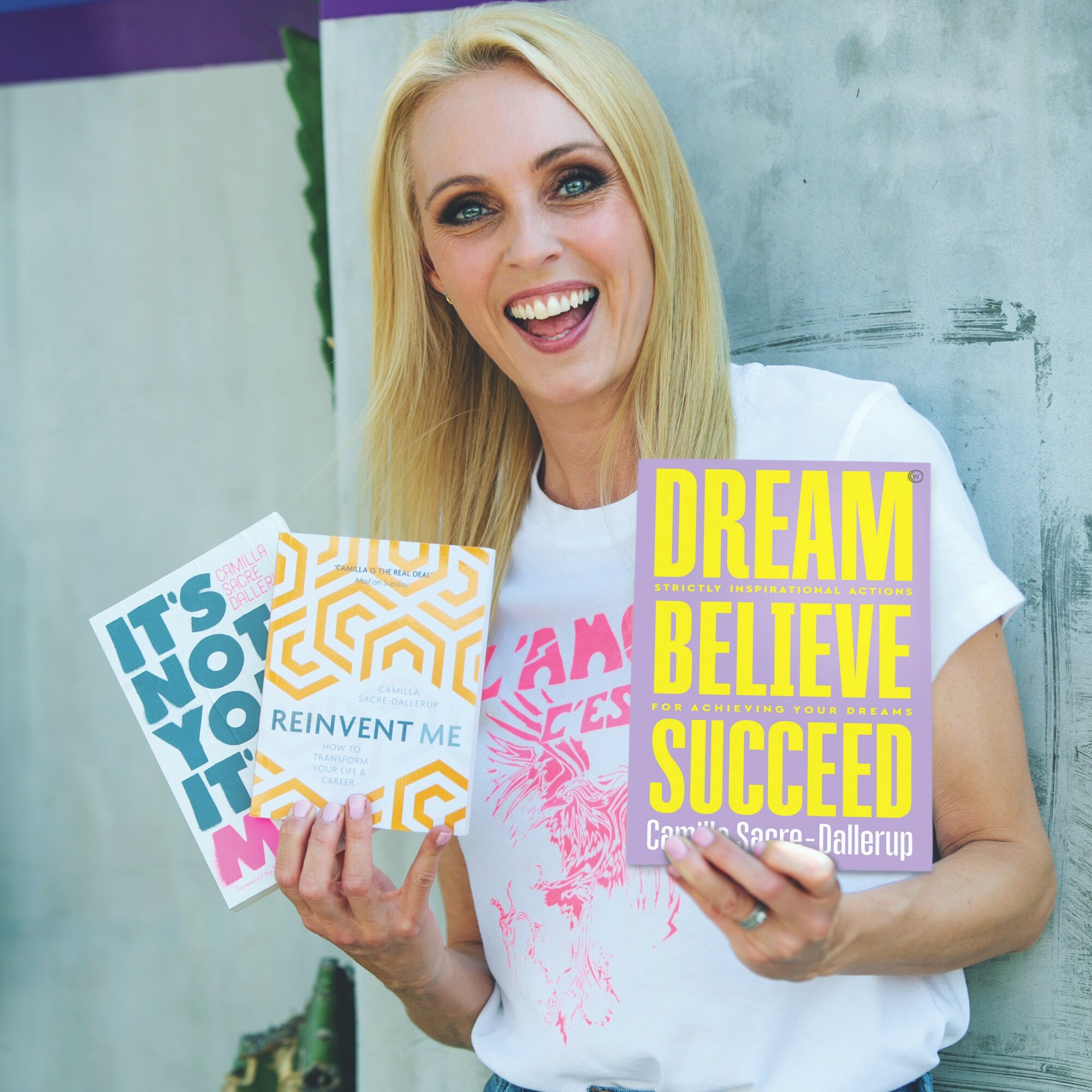 Smiling woman holding motivational signs, one reading 'DREAM BELIEVE SUCCEED' and another 'IT'S NOT YOU, IT'S ME', in front of a textured wall.