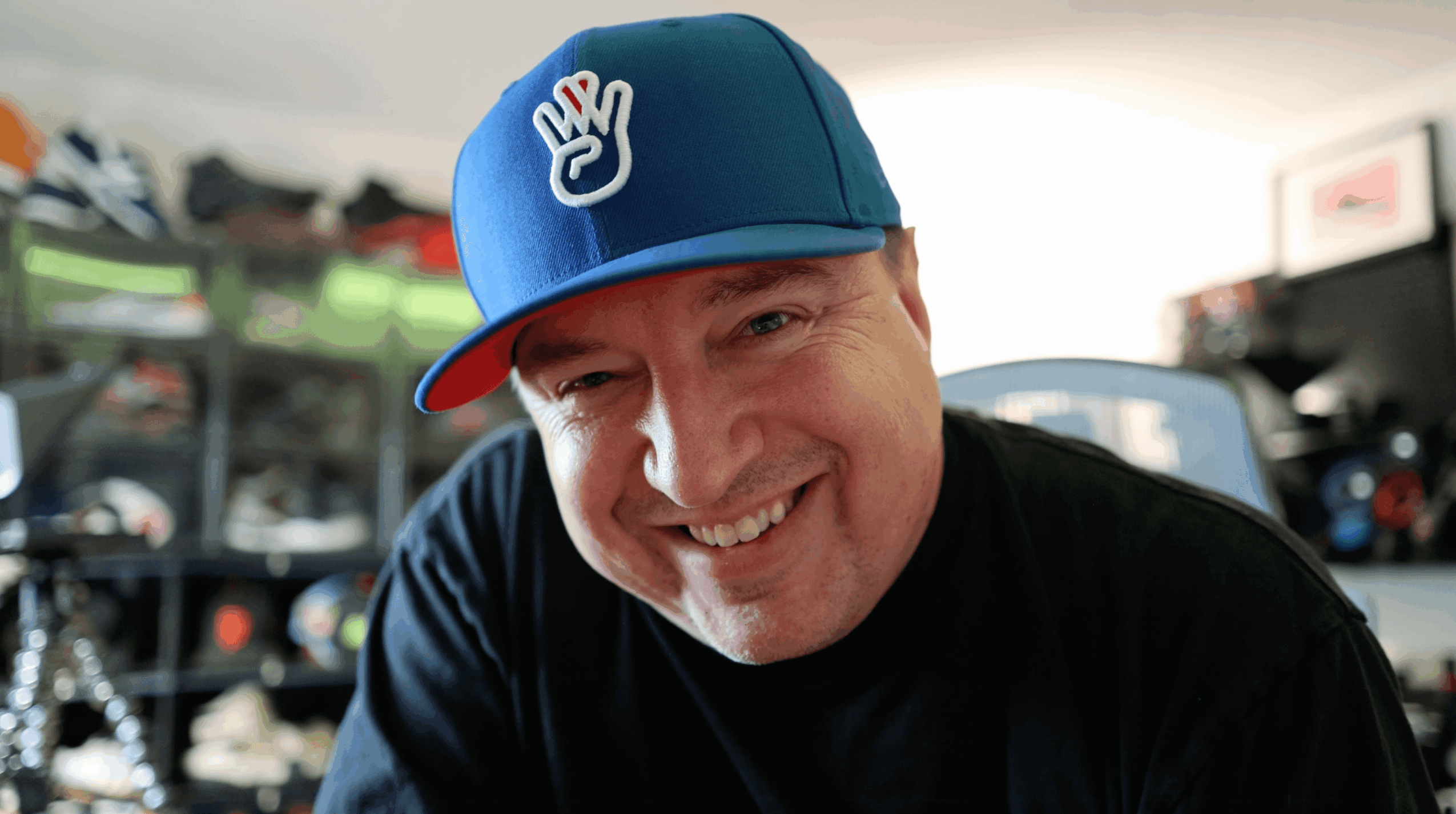 Smiling man wearing a blue cap with a hand symbol, in a cluttered indoor space.