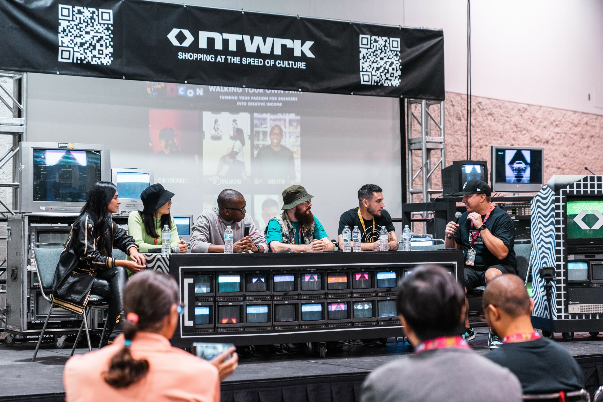 Panel of six people seated at a table on stage during a discussion or presentation, with an audience watching.