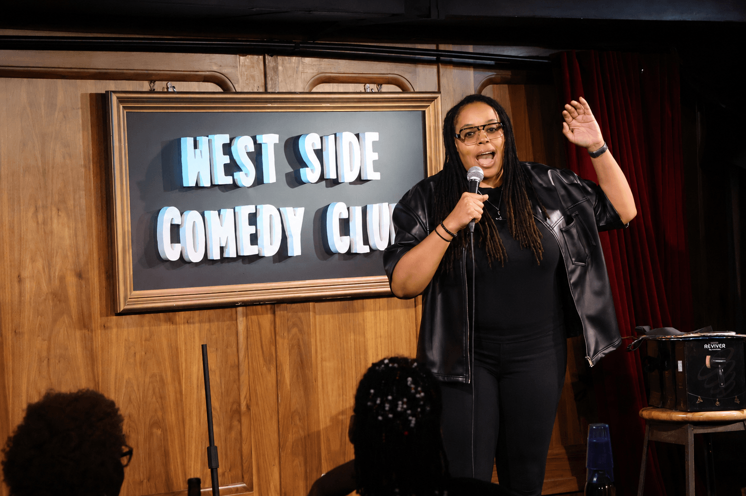 A woman performs stand-up comedy on stage at West Side Comedy Club, holding a microphone and gesturing with her hand.